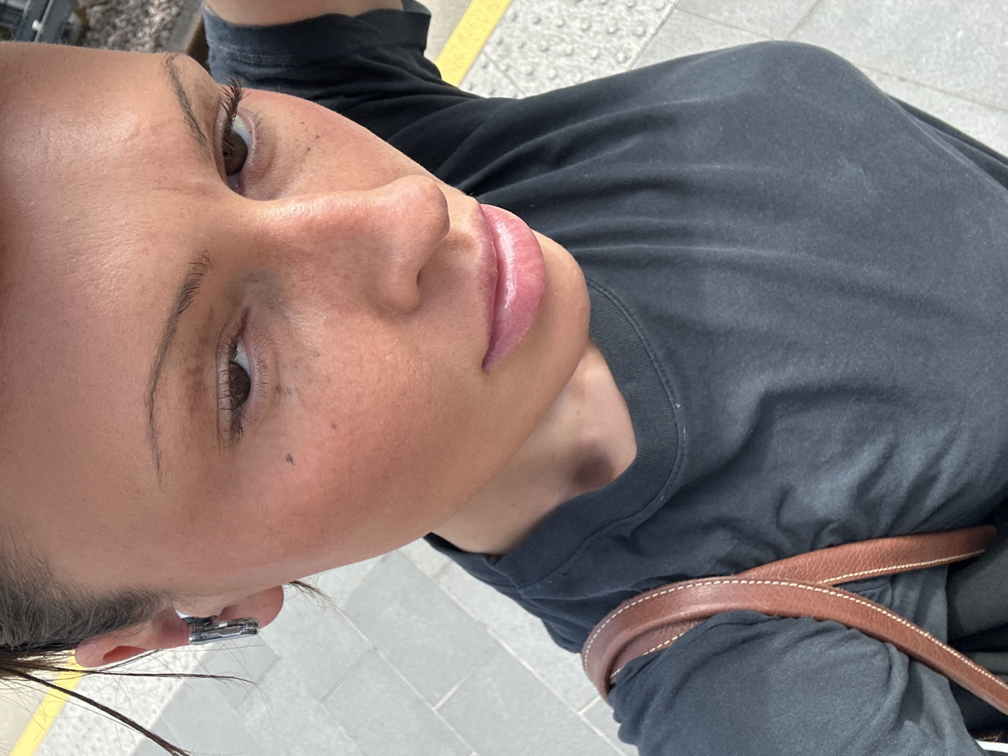 A close-up, ground-level shot focuses on a woman's face and upper torso. She is fair-skinned with brown eyes and lips that appear to have lip gloss. The woman is wearing a black t-shirt. A brown leather bag strap is visible across her torso. A silver earring is in her left ear. The ground is tiled with light gray bricks, with a small strip of yellow. Based on the tiled ground, the photograph could be taken at a London Underground station in London, UK.