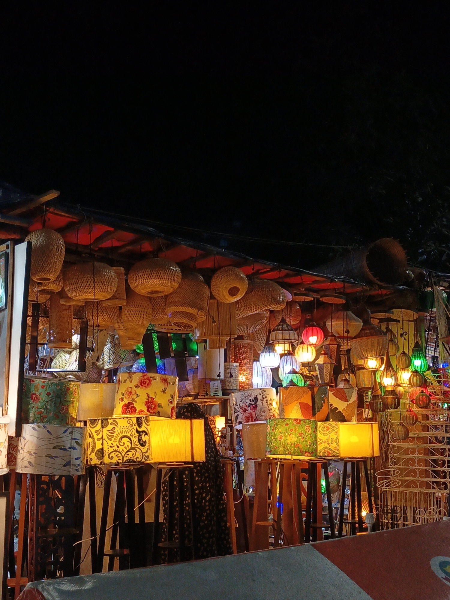 A nighttime outdoor market stall displays a variety of lamps and lanterns, illuminated by their own lights against a dark background. The stall features numerous woven bamboo pendant lamps hanging from the roof, with different shapes and sizes. Below these, a collection of table lamps with fabric shades, some adorned with floral patterns, are arranged on stands. There are also various other decorative lanterns, some with colored glass, visible throughout the display. The scene suggests a vibrant, artisanal marketplace setting.