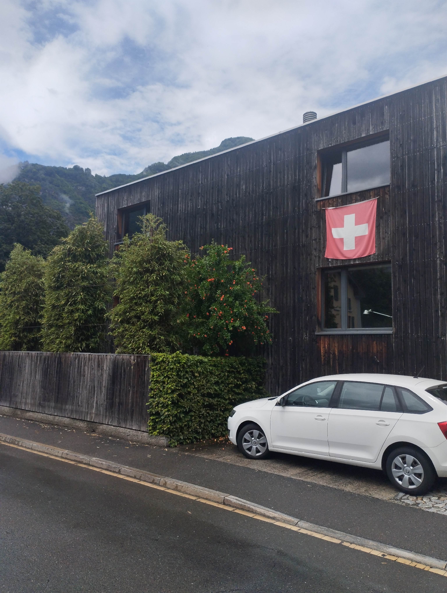 The image presents a daytime outdoor scene featuring a modern building set against a mountainous backdrop. The building's exterior is clad in dark, vertically-oriented wooden planks and features several rectangular windows. A red Swiss flag, bearing a prominent white cross, hangs from an upper-story window, indicating the location is likely Switzerland.
In the foreground, a white hatchback car, possibly a Skoda Rapid, is parked on a paved area adjacent to an asphalt road. A concrete curb and a yellow line mark the edge of the roadway.
Along the roadside, a dark wooden fence runs parallel to a neatly trimmed green hedge. Behind these, three tall, slender evergreen trees stand in a row, with a bush displaying small orange flowers positioned closer to the building on the right.
The background reveals lush green mountains, their upper sections partially veiled by low-hanging clouds or mist, suggesting an overcast or humid day. The sky above is cloudy, with diffuse light indicating it is daytime, likely late morning or afternoon. No people or specific activities are visible, contributing to a tranquil and still atmosphere.