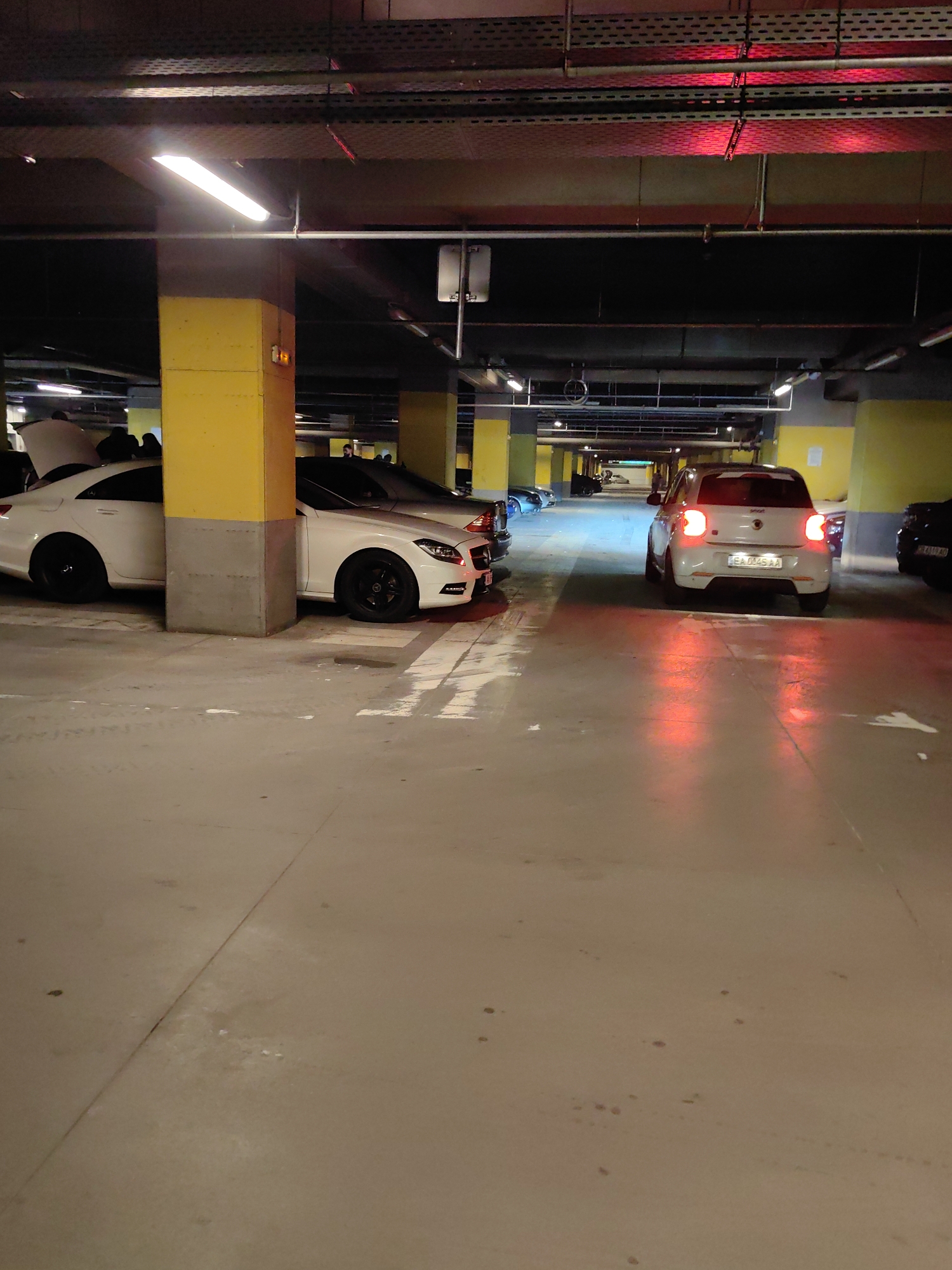 The image depicts a parking garage scene with several cars. Dominating the foreground are two white cars parked next to a yellow support pillar. One car has its trunk open, revealing several people gathered around it. Further down the garage aisle, a white Smart car with its brake lights illuminated is positioned in the center. Several other vehicles are parked further down the aisle. Overhead, the ceiling of the parking garage is exposed with lighting fixtures and cable trays. The scene is located in Sofia, Bulgaria, as suggested by the license plates of the cars.