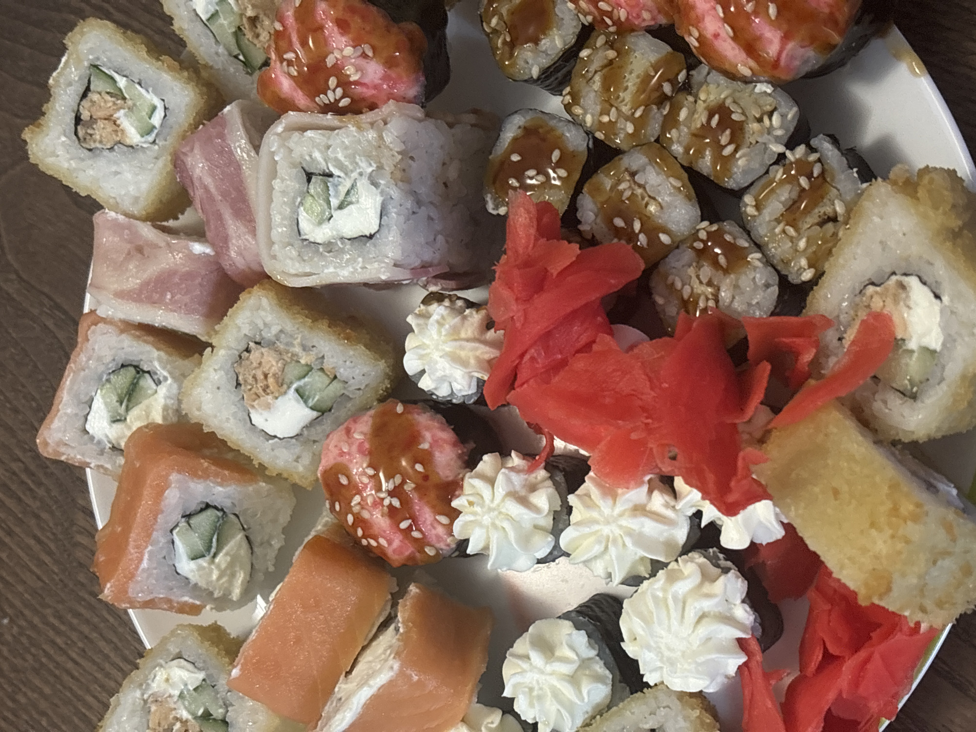 This media file displays a close-up, top-down view of a plate filled with various types of sushi and rolls. The subjects are solely food items, arranged on a white plate with a subtle green rim. The sushi includes maki rolls with visible cucumber and creamy filling, as well as some fried rolls with a golden-brown exterior. There are also salmon rolls with bright orange fish. Alongside the sushi, there are piles of pickled ginger, appearing bright pink, and dollops of white creamy sauce, possibly mayonnaise or aioli, piped in decorative swirls. The background appears to be a wooden surface, suggesting the plate is on a table. The lighting is even and bright, indicating the photo was likely taken indoors, possibly in a kitchen or dining area. There is no visible text in the image. The overall impression is a presentation of a sushi meal, ready for consumption. The arrangement suggests a home-prepared meal or a takeout order rather than a formal restaurant setting.