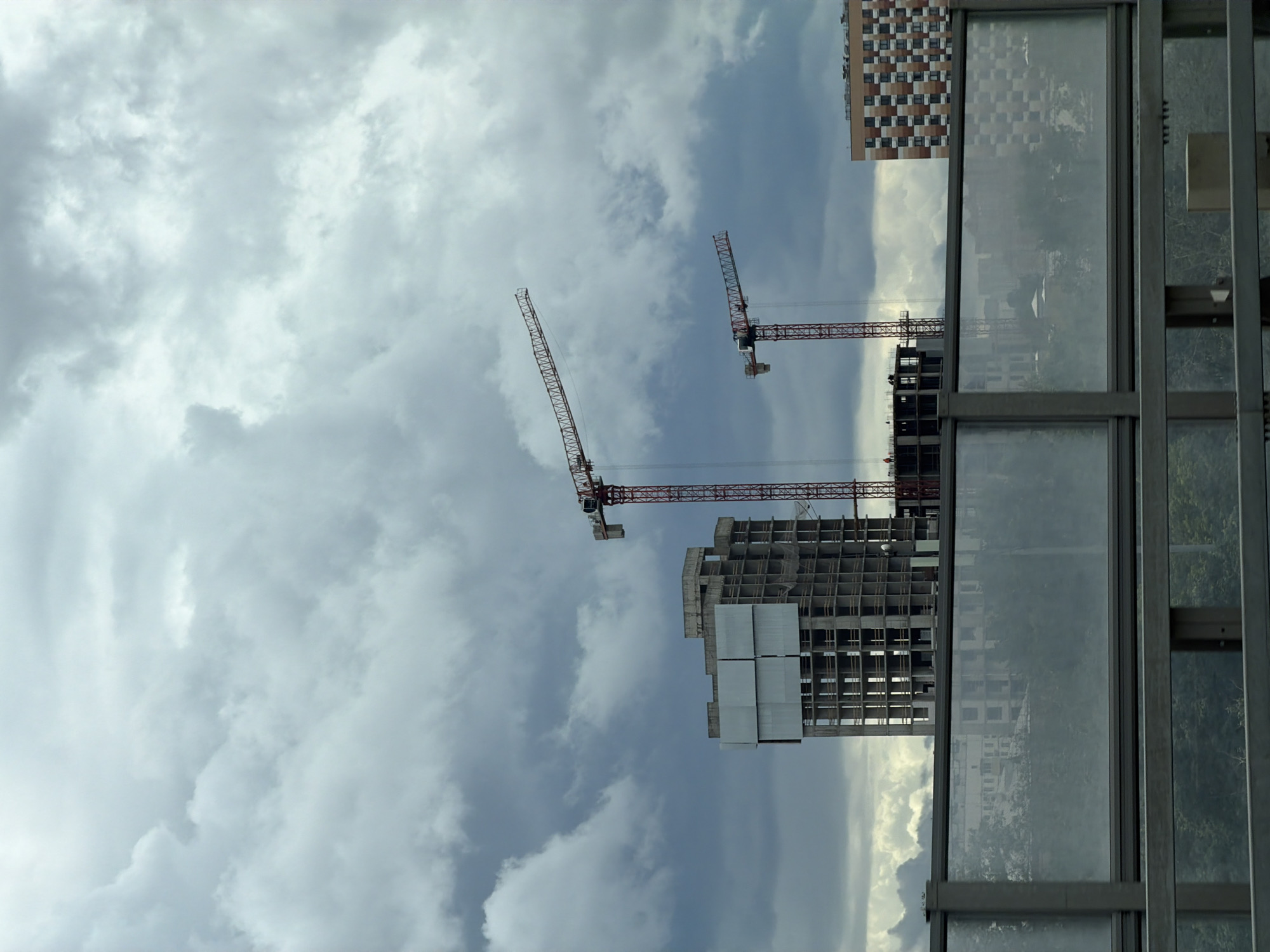 The image shows a construction site with two tower cranes and a partially built concrete building under a cloudy sky. A finished residential building with a checkered facade is visible to the right. In the foreground, there is a modern sound barrier made of glass panels and metal frames, with reflections of trees and buildings on the glass. The overall scene suggests an urban development project in Moscow, Russia, possibly near a highway or major road given the presence of the sound barrier. There are no visible people in the image, and the construction appears to be ongoing.