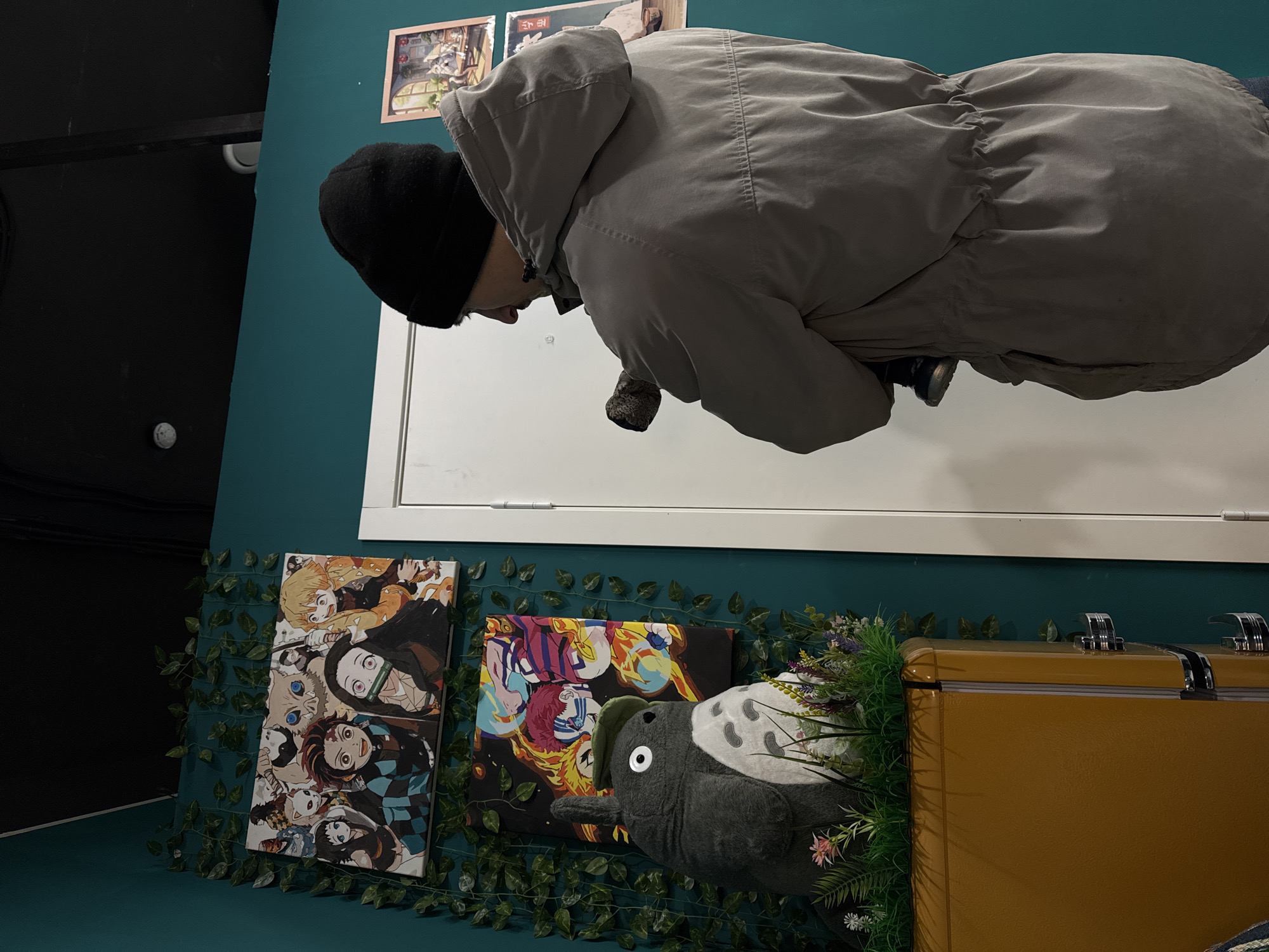 A person wearing a black beanie and a gray hooded jacket stands in a room with turquoise walls in Izhevsk, Russia. Behind the person is a white door frame. Below the door frame are two anime-themed canvas prints, a Totoro plush, faux ivy garland, and an orange cooler with metal latches. Two small framed pictures are on the wall above the person's head. The left side of the image shows a black wall and a protruding white object.