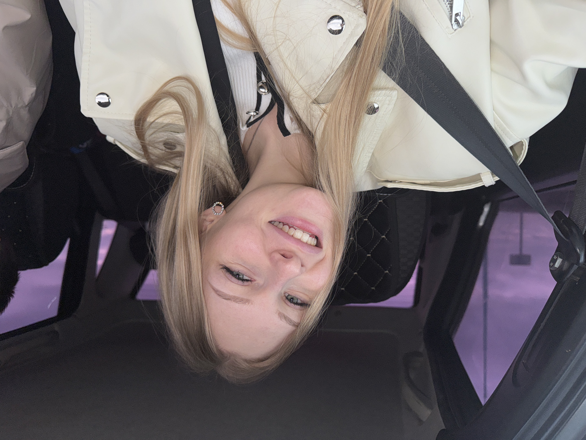 A selfie taken in a car shows a young woman with fair skin and blonde hair. She is wearing a white leather jacket and a seatbelt. The car's interior includes black leather seats with a quilted pattern. Through the car window, the sky appears purple, which suggests it is dusk or dawn. Based on the purple sky, the city of Perm, Russia, location could be accurate since it is a place in the world where these sun conditions happen.