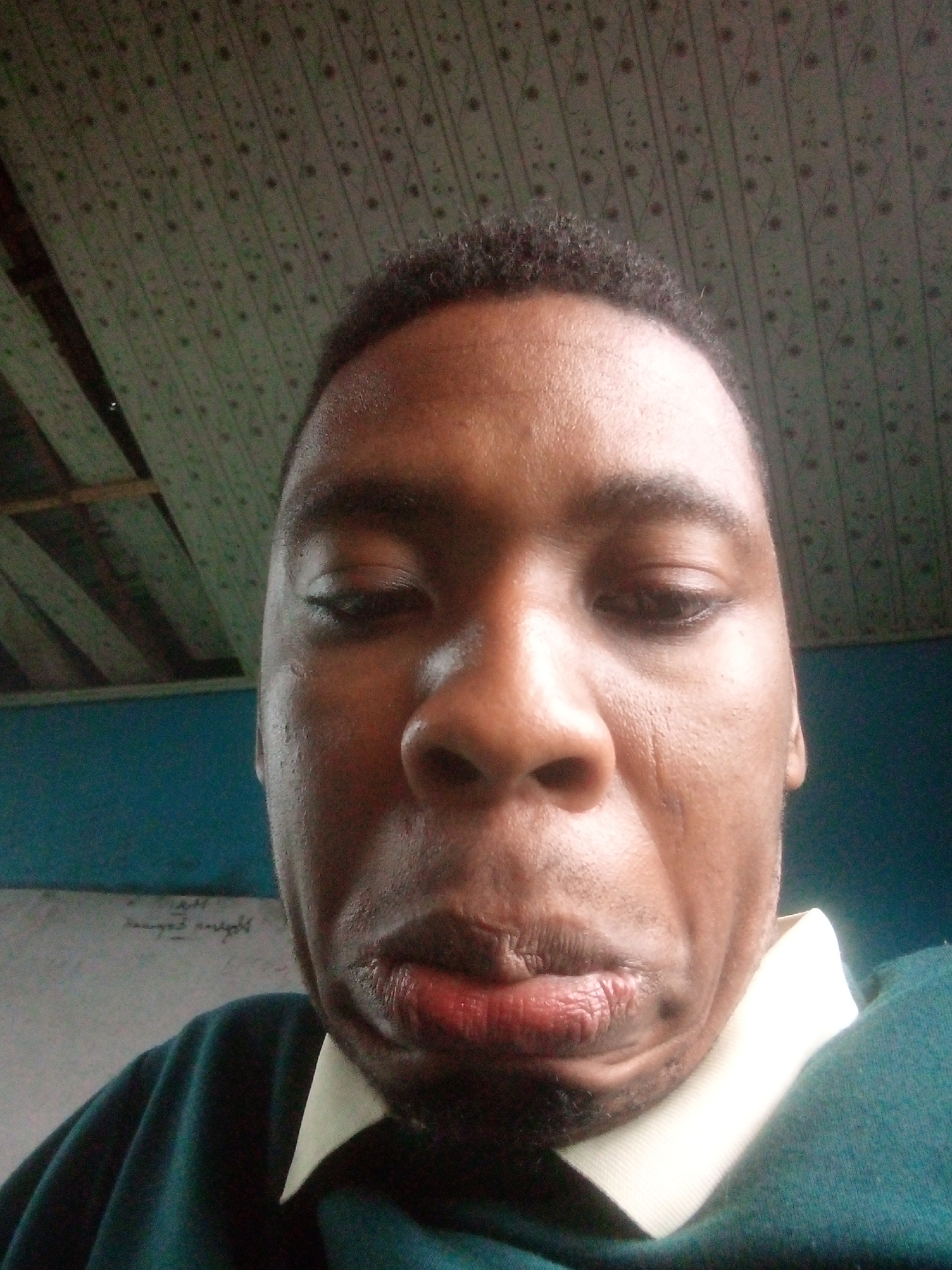 A close-up, upward-angle photograph shows a dark-skinned man with short, dark hair. He is wearing a dark green sweater over a white collared shirt. The man's expression is characterized by pursed lips and downward-looking eyes. Behind the man, there is a patterned ceiling, and a section of a blue wall. A whiteboard with writing is visible in the background. The location is Unknown, Unknown.