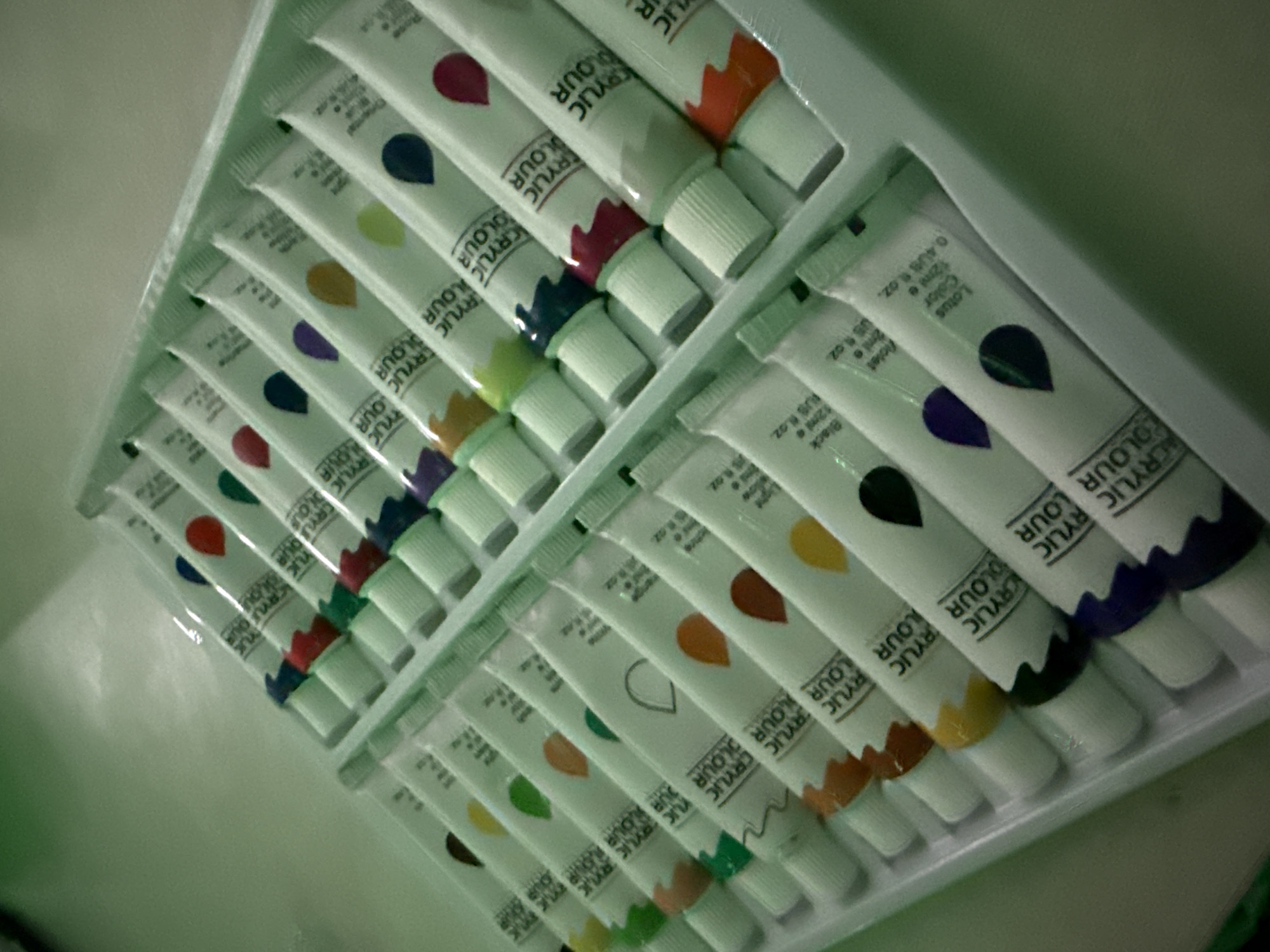 The image shows a set of acrylic paint tubes in a plastic tray. The tubes are white and labeled "ACRYLIC COLOUR." Each tube has a teardrop shape on it filled with a different color, corresponding to the color of the paint inside. The paint tubes are arranged in neat rows within the tray. The color range includes red, blue, green, yellow, orange, purple, and black. The scene is brightly lit. The location is unknown. There are no people present in the image.