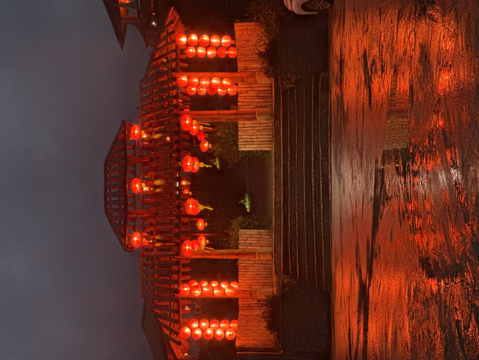 The scene depicts a brightly illuminated Chinese-style gateway at night, located in Ban Mai Saha Sampan, Thailand. The gateway is constructed from dark wood or bamboo, featuring multiple tiered roofs. Numerous red lanterns are suspended from the structure, casting a dominant crimson glow over the entire area. Vertical columns of illuminated red lanterns flank the entrance, with additional individual lanterns hanging from the roof eaves.
A series of dark, multi-tiered stone steps leads up to the gateway. In the immediate foreground, a wet, dark paved surface (likely asphalt or concrete) reflects the intense red light from the lanterns, creating blurred streaks and pools of color. The reflections indicate recent precipitation. Beyond the gateway's archway, a dark, indistinct path recedes into the background, with a faint green light source visible further along the path. To the far right of the paved area, a portion of a white passenger vehicle, specifically its front passenger side wheel and fender, is visible. The sky above is dark, confirming nighttime conditions. No individuals are present within the frame.