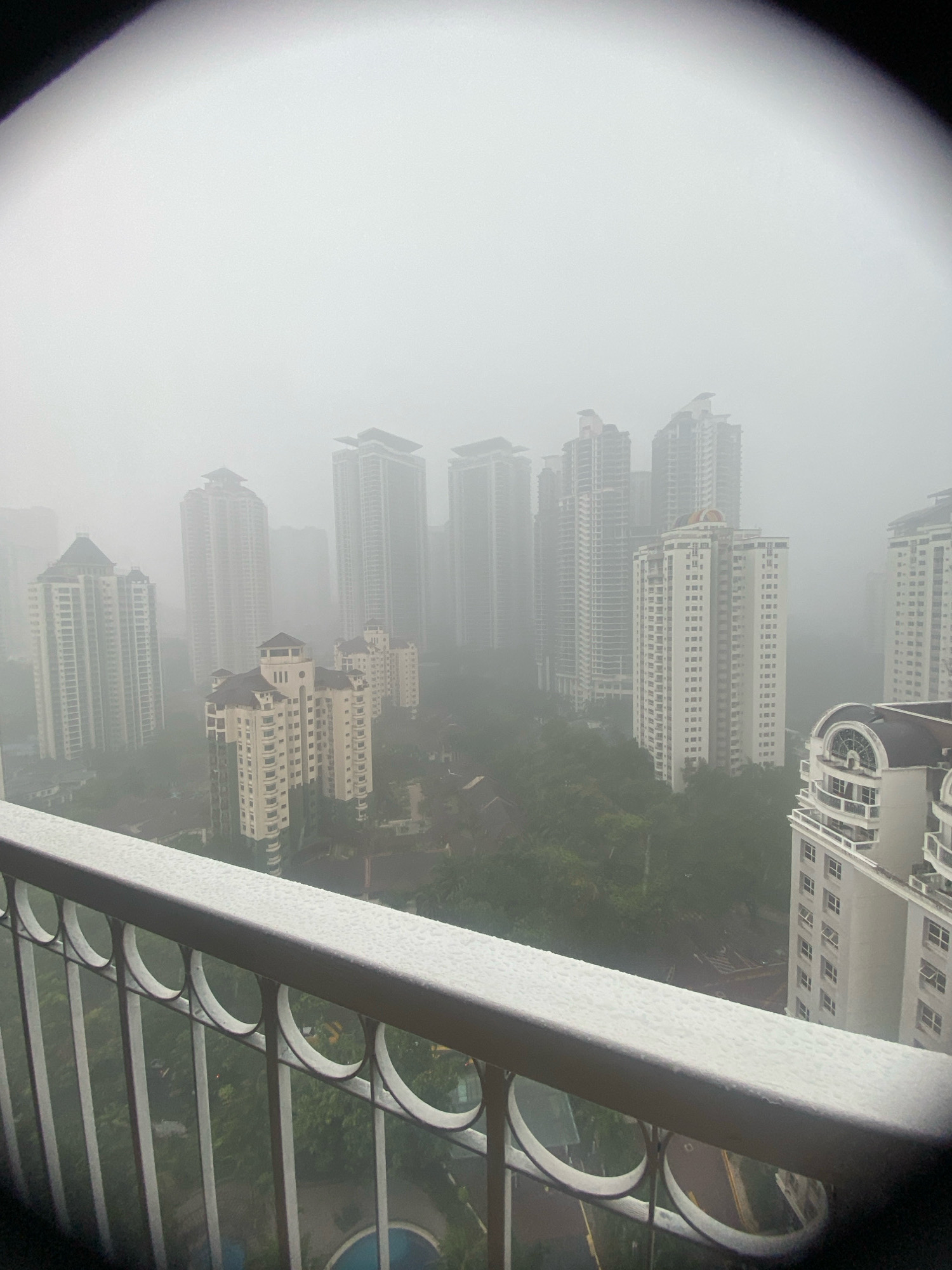 The image is a high-angle, wide shot of a city skyline obscured by heavy fog or mist, likely taken from a residential building in Kuala Lumpur, Malaysia, given the architectural style of the buildings and the lush greenery visible. In the foreground, a white balcony railing with decorative circular metalwork is visible, wet with what appears to be rain or condensation. Beyond the railing, numerous high-rise apartment buildings of varying heights and architectural designs populate the landscape. The buildings are mostly light-colored, with many featuring balconies and distinct rooflines. Some buildings are modern glass and steel structures, while others have more ornate facades with protruding sections and distinct tops. The dense fog significantly reduces visibility, creating a hazy, almost ethereal atmosphere and obscuring details of the farthest structures. Below the buildings, patches of dense green trees and some infrastructure like a swimming pool are faintly discernible through the mist. The overall impression is one of a city enveloped in a thick layer of atmospheric moisture, creating a somber and somewhat mysterious mood. No people or active events are depicted in the scene.