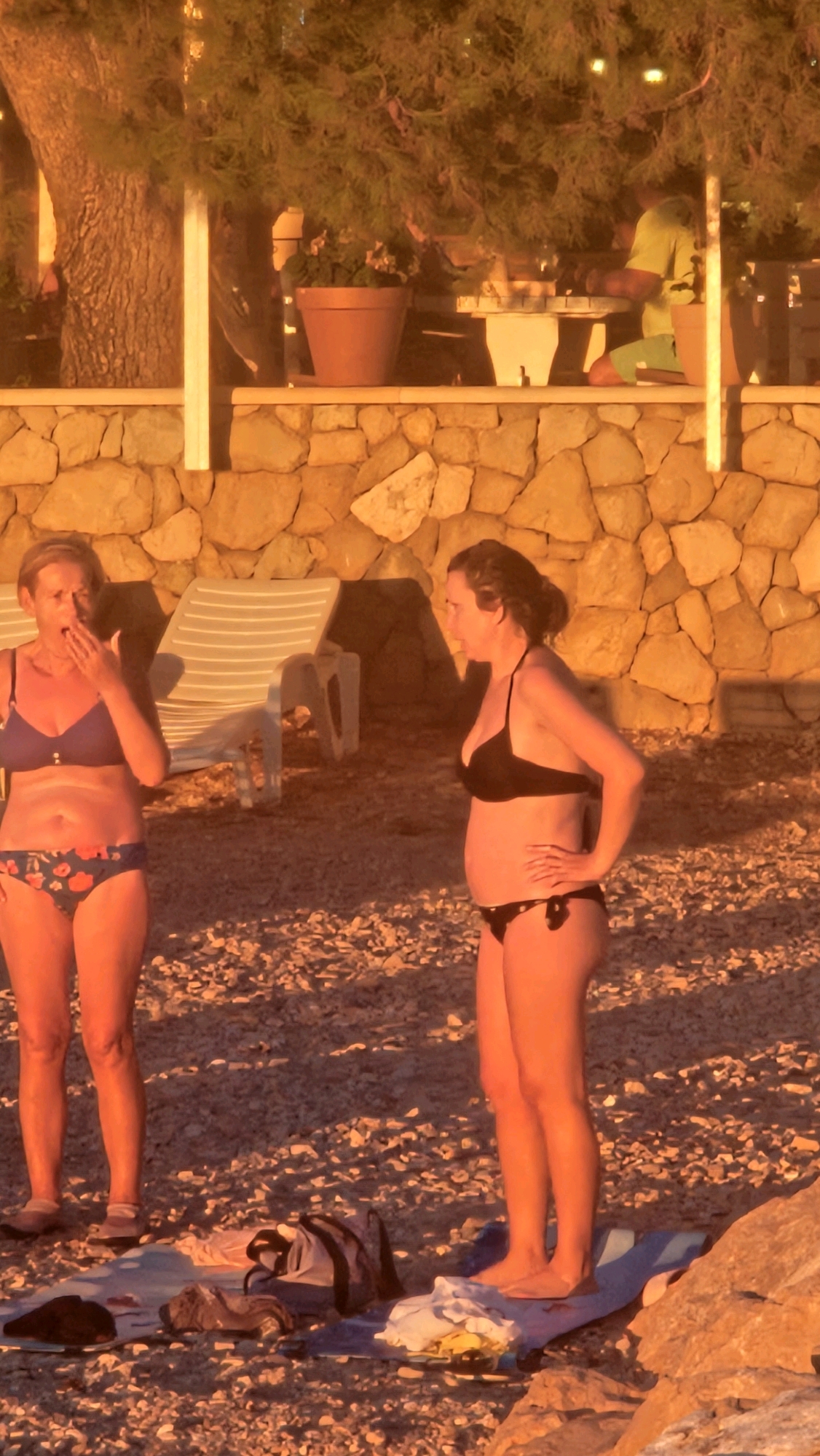 Two women in bikinis are standing on a pebble beach. The woman on the left is wearing a blue bikini with a floral pattern on the bottoms, and she is covering her mouth with her hand, as if yawning or speaking. The woman on the right is wearing a black bikini and has her hand on her hip. Behind them, there are lounge chairs and a stone wall. In the background, a person in a yellow shirt and green shorts is sitting at a table. The scene is illuminated by warm, golden light, suggesting it is either sunrise or sunset. The location is Općina Tučepi, Croatia.