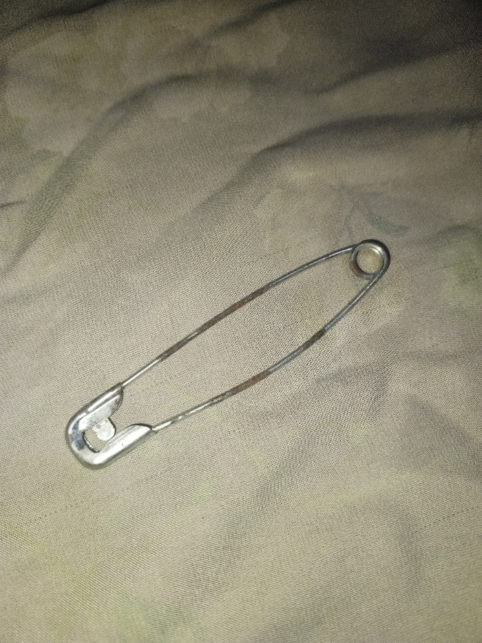 A single, open metallic safety pin lies diagonally on a textured, desaturated green fabric surface in Hasanpur, India. The safety pin's silver-toned body exhibits segments of brown-orange corrosion along its wire frame. The clasp mechanism is oriented towards the lower-left, while the coiled spring mechanism is towards the upper-right. The underlying fabric displays a visible weave pattern and subtle creasing. No individuals, actions, or interactions are present.