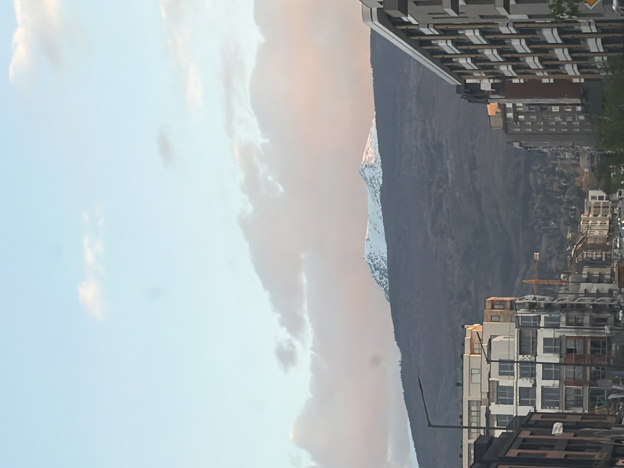 This image, presented with a 90-degree clockwise rotation, depicts a panoramic view of a city, identified as Sofia, Bulgaria, framed by a prominent mountain range, likely Vitosha. The scene suggests either sunrise or sunset, characterized by a light blue sky transitioning into soft pink and orange cloud formations across the upper and central portions of the frame.
In the foreground, on what would be the right side of a correctly oriented image, a series of modern, multi-story residential buildings with balconies and diverse architectural details are visible. Some structures appear to be under construction or newly built. Further into the mid-ground, beneath the mountain, more city buildings and a distinct yellow construction crane can be seen.
The dominant feature is the expansive mountain range, its dark, forested slopes occupying the central and lower left areas. A majestic, snow-capped peak rises distinctively, illuminated by the warm, low-angle sunlight. The combination of the urban environment and the natural landscape creates a striking contrast. There are no discernible human activities taking place, nor is any legible text visible within the frame.