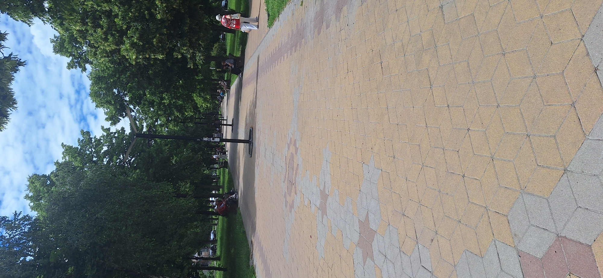 The image depicts a sunlit park path in Bila Tserkva, Ukraine. The path is paved with interlocking hexagonal and diamond-shaped stones in shades of yellow, gray, and reddish-brown, creating a geometric pattern. Mature trees with lush green foliage line both sides of the path, forming a canopy overhead. Several lampposts with a modern design are spaced along the path.
In the foreground, the patterned paving is clearly visible. Further along the path, there are several people. On the left side, three individuals are seated on a bench, conversing. On the right side, closer to the foreground, a person wearing a red and white shirt and white pants is standing and facing away from the camera. Further down the path, other individuals are seen sitting on benches or walking. In the background, parked cars are visible on a street bordering the park, and buildings can be seen in the distance. The sky is blue with scattered white clouds, suggesting fair weather. The overall scene conveys a tranquil atmosphere in a public park setting.