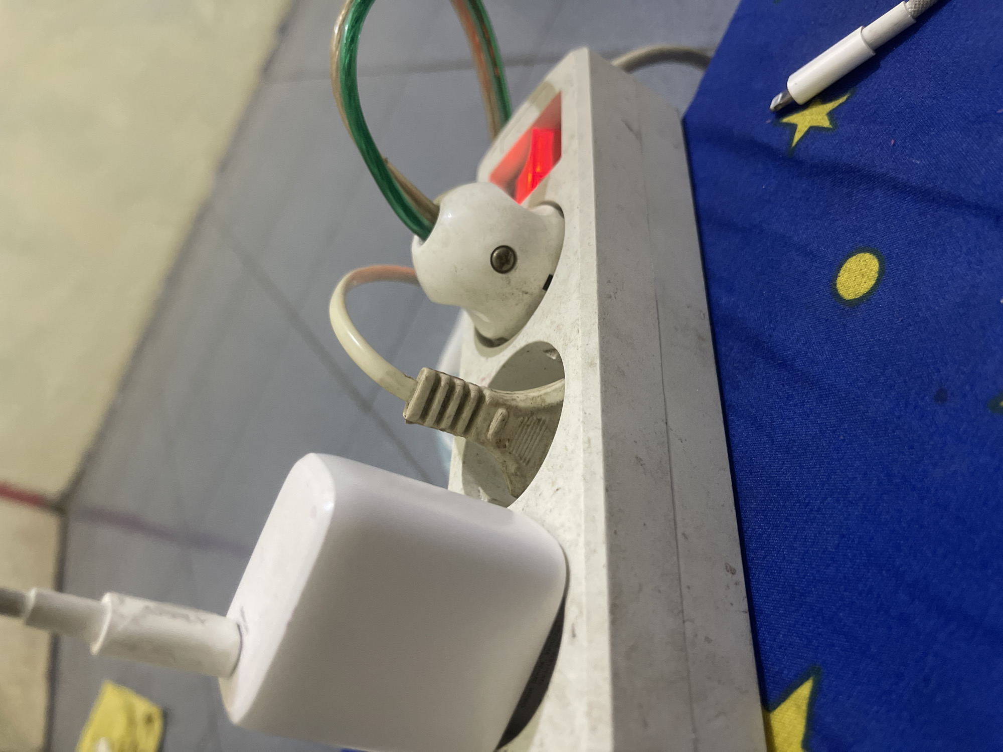 A white power strip is plugged into a wall socket, with a white phone charger and two other plugs inserted into its outlets. One of these plugs has a red indicator light, suggesting it is receiving power. The power strip and plugs are situated on a blue surface adorned with yellow stars and circles, possibly a fabric. In the background, a tiled floor and a wall are visible. The scene appears to be indoors, likely in a residential setting within Anggaswangi, Indonesia.