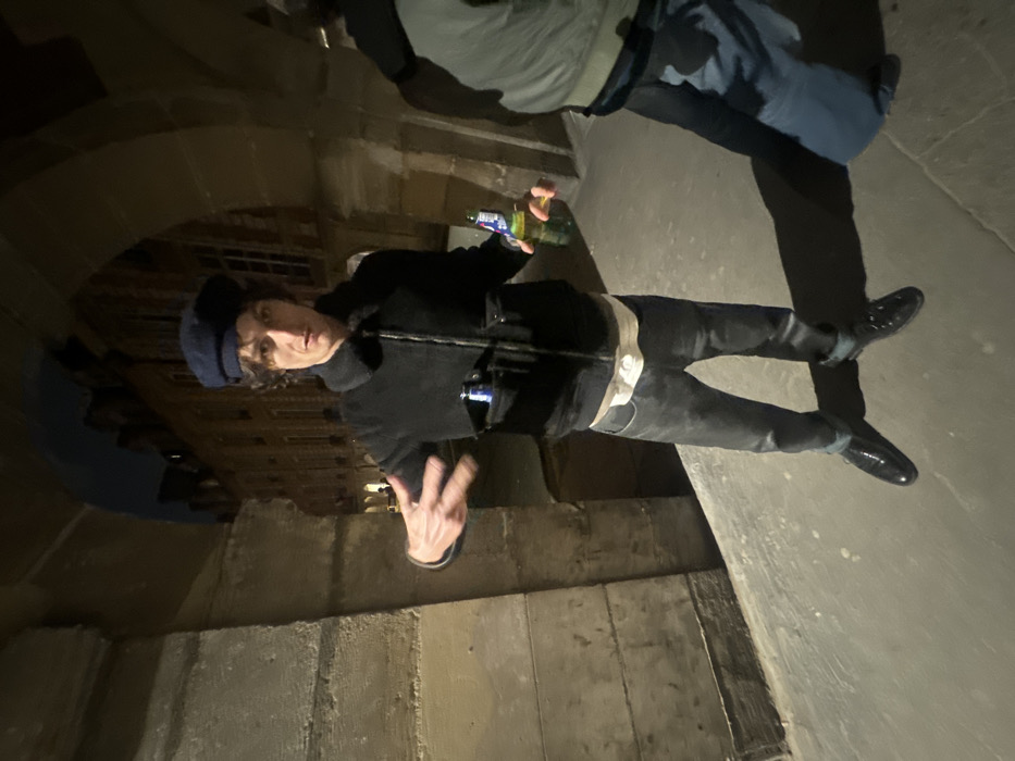 A person wearing a black jacket, dark pants, and shiny black shoes stands in a dimly lit area. The person also wears a navy-colored cap and holds a green bottle in one hand, gesturing with the other. Another person, partially visible at the top of the frame, wears a light-colored jacket and blue pants. The background includes architectural features with stonework and what appears to be a window or opening.