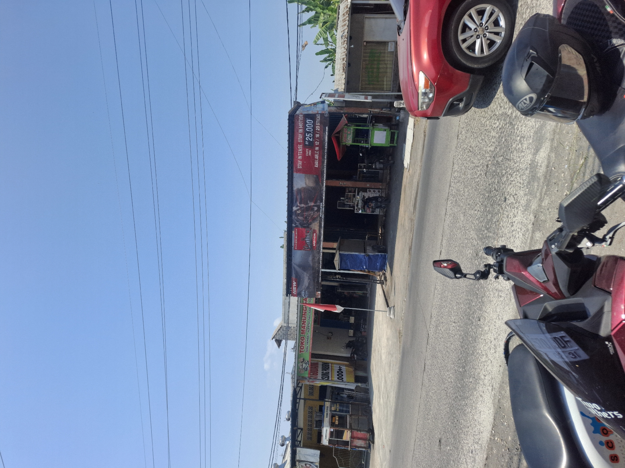 The image depicts a street scene in Sumberwudi, Indonesia, on a clear, sunny day. In the foreground, the front portion of a dark red motorcycle with a black helmet resting on its seat is visible. To the right, a red car is parked on the side of the road, facing left. The background features a row of buildings with storefronts, including a shop with a prominent banner advertising "STAY INTENSE. STAY IN MOTION" and "Rp 25,000" with availability in various stick counts. A red and white Indonesian flag is planted in front of one of the stores. Several other storefronts are visible, with various signs and advertisements, including one that reads "TOKO MANUNGGAL" and mentions "JUAL SANDAL, SEPATU, TAS, SABU TO.". A green food cart with the text "PUTUT SOY" is positioned in front of one of the stores. The road surface is asphalt, and power lines crisscross the sky. Trees, including banana trees, are visible on the far right of the image.