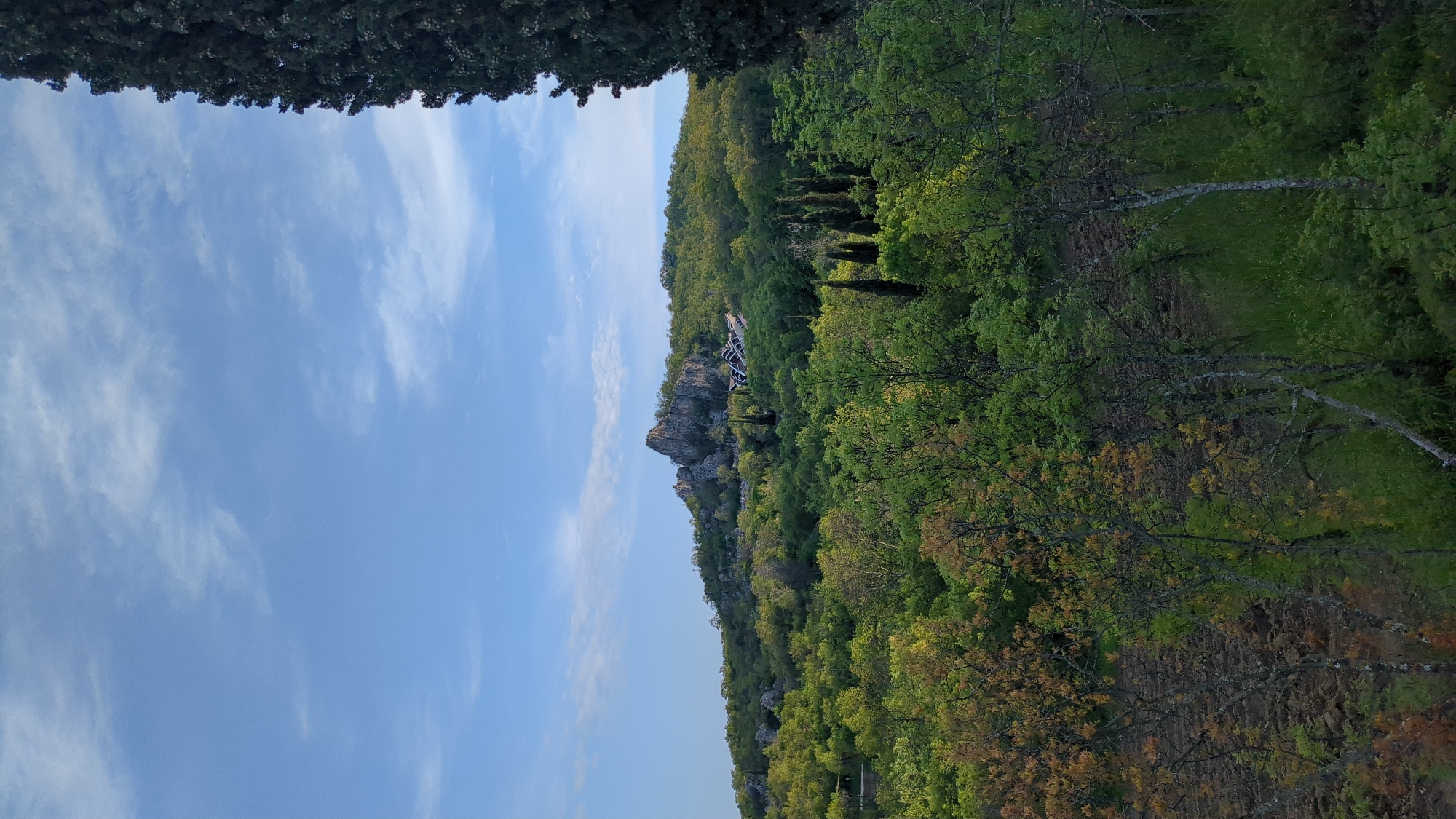 The image depicts a landscape scene in Unknown, Unknown, featuring a clear blue sky with scattered clouds in the upper left portion. Below the sky, a tree line defines the top edge of a densely vegetated hillside. The hillside is covered with various shades of green foliage, interspersed with trees showing hints of autumn colors. A rocky outcrop is visible, featuring a structure (possibly a building or architectural element) on top. A stand of tall, thin trees stands further up the slope. In the foreground, a path or trail is visible, leading into the wooded area. A dark mass of trees is at the top.