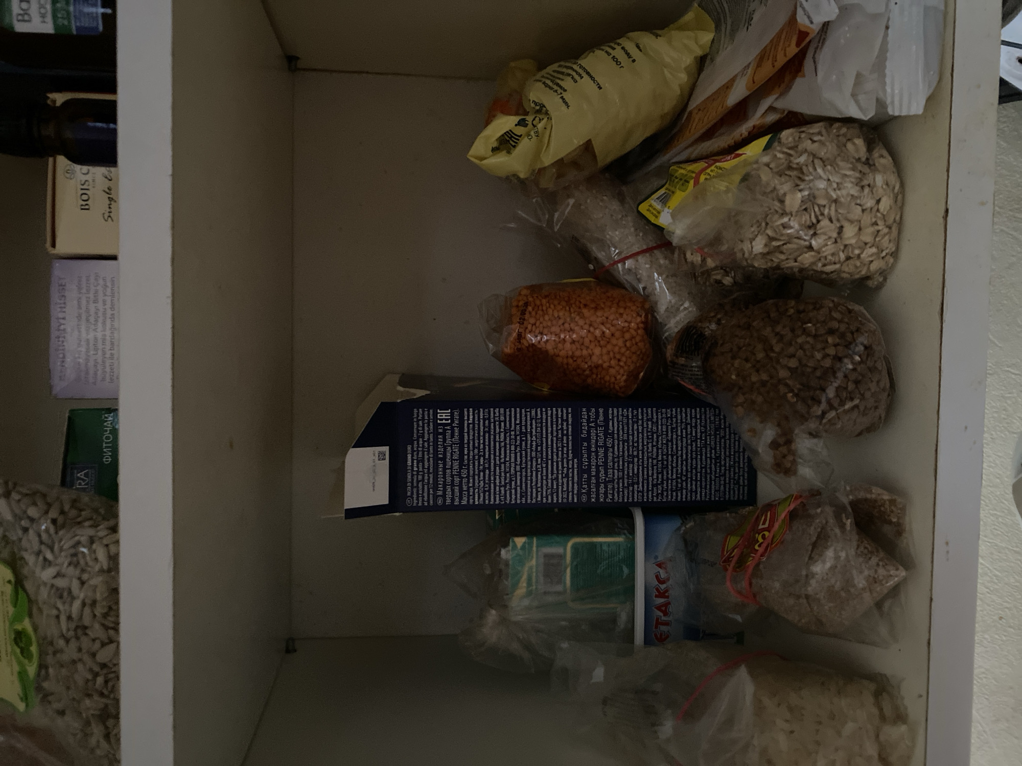 The image depicts a shelf in a pantry, likely in Sokhumi, Abkhazia, as suggested by some of the visible text. The shelf is organized with various food items, primarily dry goods, stored in plastic bags and boxes.
In the foreground, from left to right, are several clear plastic bags containing foodstuffs. The leftmost bag appears to hold a light-colored grain, possibly rice. Next to it, a bag contains a darker, granular substance, possibly buckwheat or another type of grain. A similar bag is positioned to the right of this, containing what also appears to be a grain. Further to the right, a bag holds rolled oats.
Behind these items, on a higher shelf, are more prominently displayed products. A large blue and white box with the word "ETAKCA" is visible. To the left of this box, a white and blue cardboard box is partially obscured. Behind these, a red-lidded container and a yellow bag with an illegible label are present.
On the top shelf, there are several smaller items. A clear plastic bag filled with sunflower seeds is on the far left. To its right, a green box with the Cyrillic word "ФИТОЧАЙ" (which translates to "herbal tea") is visible. Next to this is a white box with text in a foreign language, and a small dark bottle.
The lighting suggests an indoor environment, likely a domestic kitchen or pantry. There are no people or animals present in the image. The overall impression is one of storage and organization of food supplies.