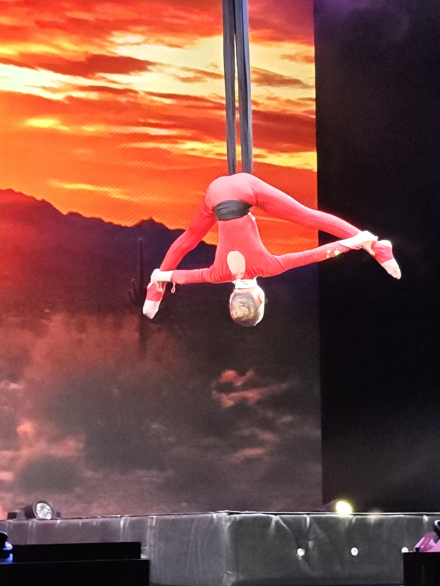 A female aerialist in a red jumpsuit performs suspended from fabric straps. She is bent over backwards with her legs split wide. The performance is taking place indoors on a stage with a screen displaying a desert-like landscape. Based on the information I have, I cannot confirm if this image was taken in Yekaterinburg, Russia.
