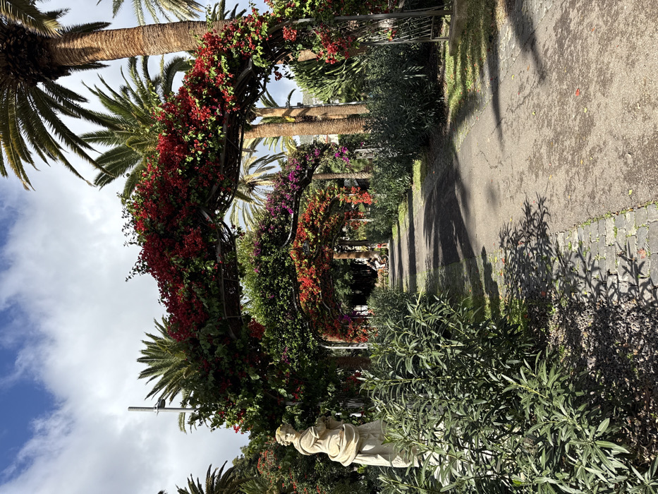 The image displays a pathway within a landscaped garden in Santa Cruz De Tenerife, Spain, under a partially cloudy sky. In the immediate foreground, to the left, a light-colored classical-style female statue, appearing to be carved from stone, stands amidst green shrubs. The pathway, constructed of asphalt with a cobblestone edge on the left, extends into the distance, flanked by dense green foliage. A series of dark-colored metal archways, heavily adorned with vibrant red and some purple bougainvillea and green vines, span the path, creating a tunnel effect. Numerous tall palm trees with brown trunks and green fronds are interspersed throughout the scene. A solitary individual on a light-colored bicycle or scooter is visible on the pathway in the mid-distance, moving away from the viewer. The background consists of further dense vegetation and additional palm trees.