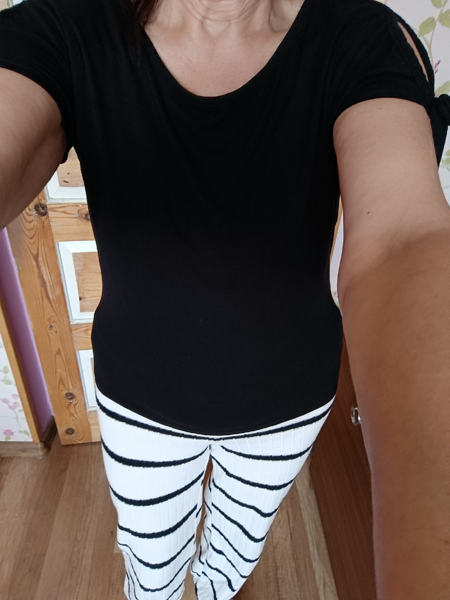 The image shows a person wearing a black t-shirt with cut-out sleeves tied with bows and white pants with black horizontal stripes. The person's arms and torso are visible from the shoulders down to the knees. The background includes a wooden door and wall with wallpaper. The scene appears to be indoors.