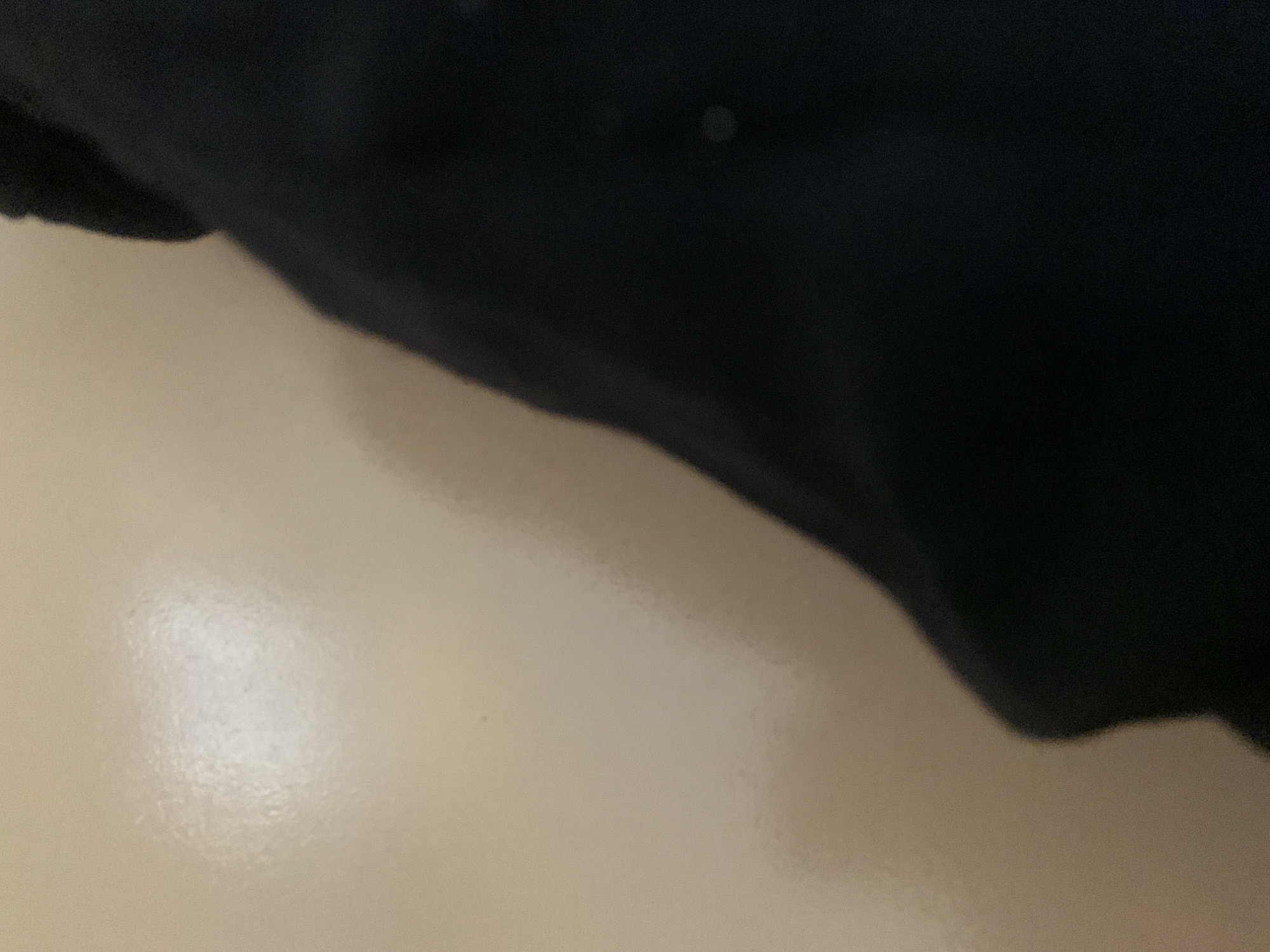 The image shows a close-up view of a beige surface with a sheen, possibly a wall or a piece of furniture. A dark, possibly black, fabric is draped over the right side of the frame, obscuring a portion of the beige surface. The fabric appears to be soft and has some wrinkles. There is a blurred, bright light source reflecting off the beige surface in the upper left quadrant of the image. The scene is devoid of any identifiable landmarks or context that would suggest a specific location within Jauro Yinu, Nigeria. There are no people, actions, or interactions visible in the image. The overall impression is one of abstract texture and light play, offering no narrative or specific environmental details.