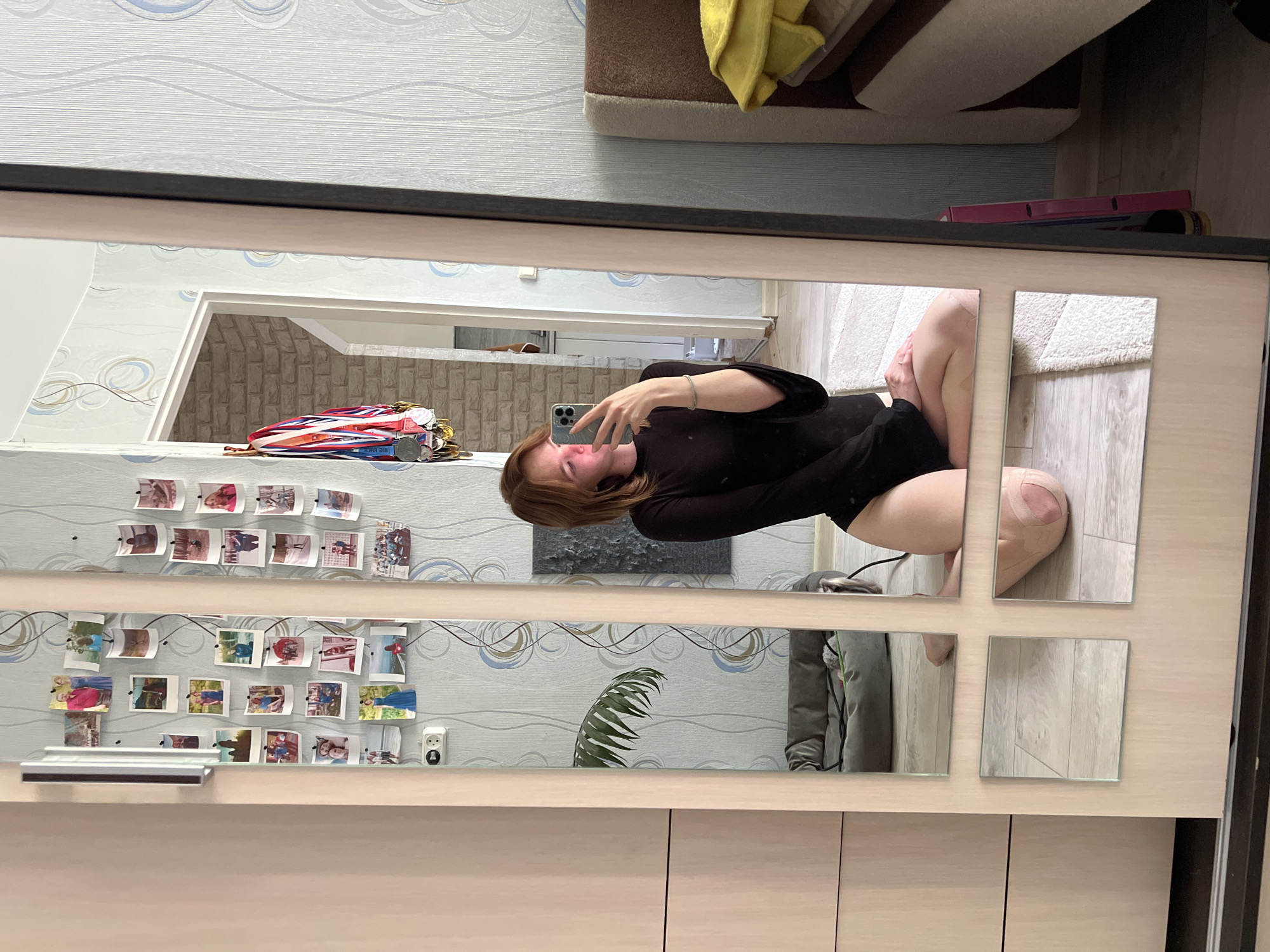 This image is a mirror selfie taken by a person in an indoor setting, likely a home in Chaikovsky, Russia. The perspective is from the mirror, showing the person holding a smartphone to capture their reflection.
The central subject is a young person with reddish-brown hair, wearing a black long-sleeved top. They are seen kneeling or squatting on a light-colored wooden floor, with visible bandages or tape on both knees. The smartphone they are holding appears to be a modern model, possibly an iPhone, and its screen shows the camera interface.
The room reflected in the mirror is personalized and lived-in. The wall behind the person is decorated with numerous small printed photographs or polaroids arranged in a collage, interspersed with a light-toned wallpaper featuring wavy patterns. A white electrical power outlet is also visible on this wall. To the reflected left of the person, a collection of colorful keychains and medals rests on a surface, one of which visibly says "BIG RICE" (mirrored). A dark, textured artwork hangs on the wall behind the person's head.
Other reflected objects include a part of a sofa or bed with a yellow blanket/cushion in the upper right corner, and a white shaggy rug covering a portion of the wooden floor. A large green, leafy plant is also reflected, indicating its presence in the room. The large mirror itself is framed and appears to be part of a wardrobe or a standing unit, with smaller square mirror panels integrated into its lower right section. Below the mirror, a light-colored wooden wardrobe or cabinet is visible. Above the mirror's reflection, a pink binder or folder is placed on a shelf. The indoor lighting suggests daytime or a well-lit interior.
