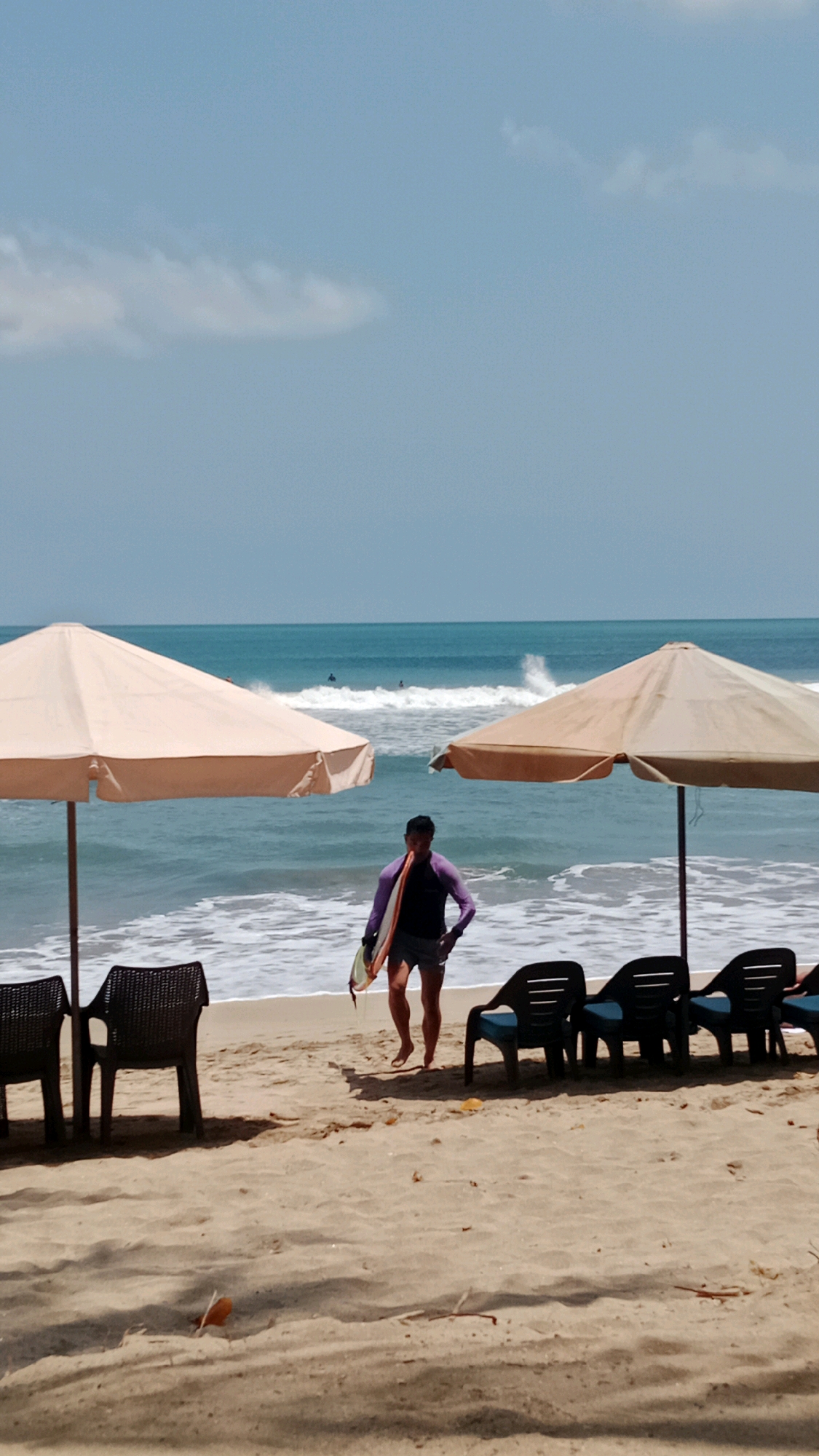 This is a full, vertical shot of a sandy beach in Kuta, Indonesia, on a sunny day with a light breeze. In the foreground, two large, beige beach umbrellas provide shade over empty plastic chairs. A person is walking from the ocean towards the shore, carrying a surfboard. The person is wearing a purple long-sleeved rash guard and dark shorts. The ocean is visible in the midground, with gentle waves breaking on the shore and several surfers in the distance. The sky is a pale blue with a few wispy clouds. The sand is light brown and appears dry.