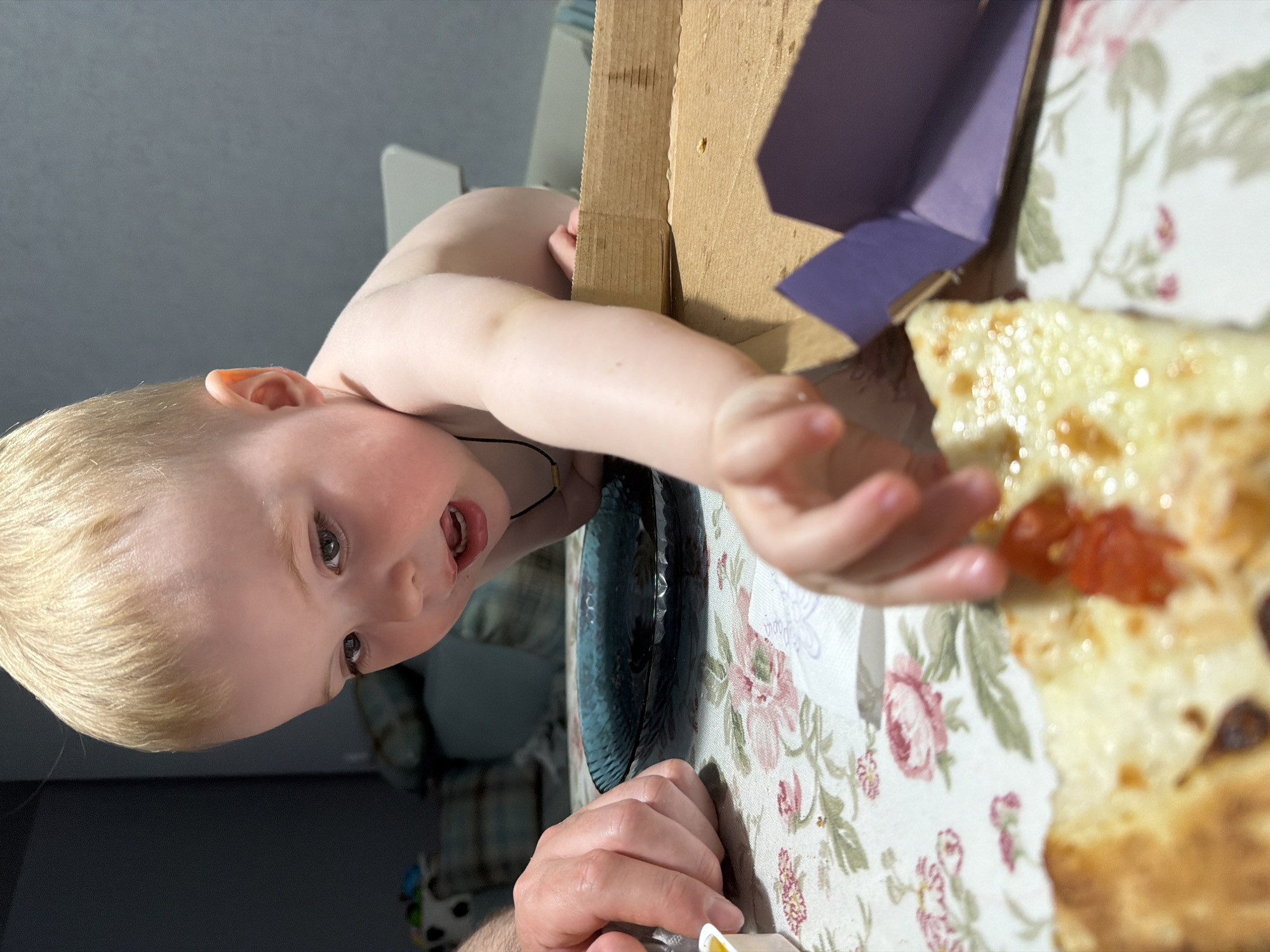 A young, fair-skinned child with blond hair reaches for a slice of pizza from a pizza box on a table covered with a floral-patterned tablecloth. A blue textured plate sits near the pizza box. An adult's hand is visible near the bottom of the frame, holding a small white object. The setting is indoors, possibly a home. The location in the city of Votkinsk, Russia, cannot be determined from the image.