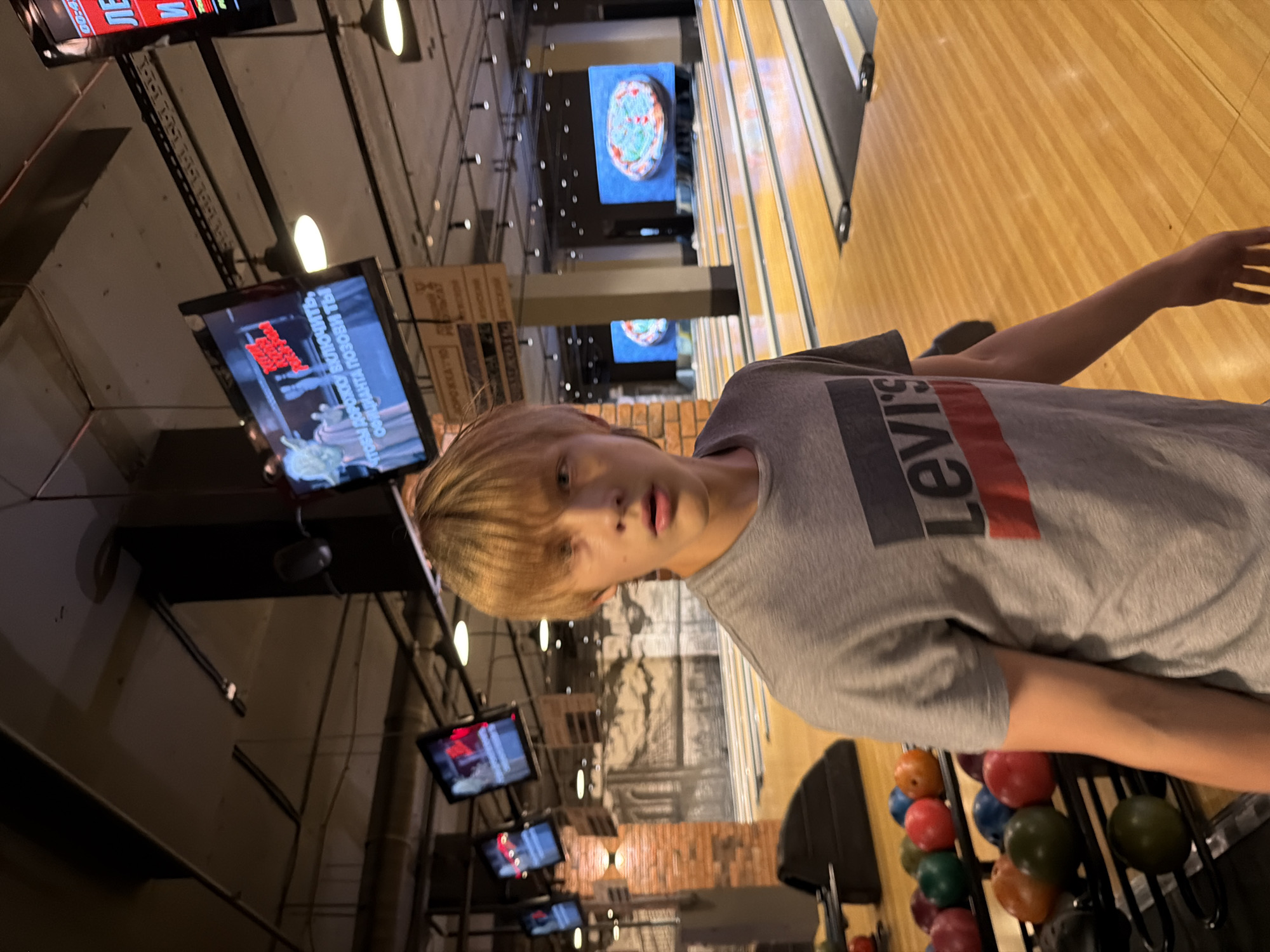 A young person with blonde hair, wearing a gray t-shirt with a Levi's logo, stands in a bowling alley. They are looking towards the camera with a neutral expression. The bowling alley features wooden lanes, and bowling balls of various colors are visible on racks to the right of the frame. Several screens are mounted on the wall in the background, displaying images and text. One screen shows a close-up of a pizza. The lighting suggests it is indoors. The provided location context is Izhevsk, Russia.