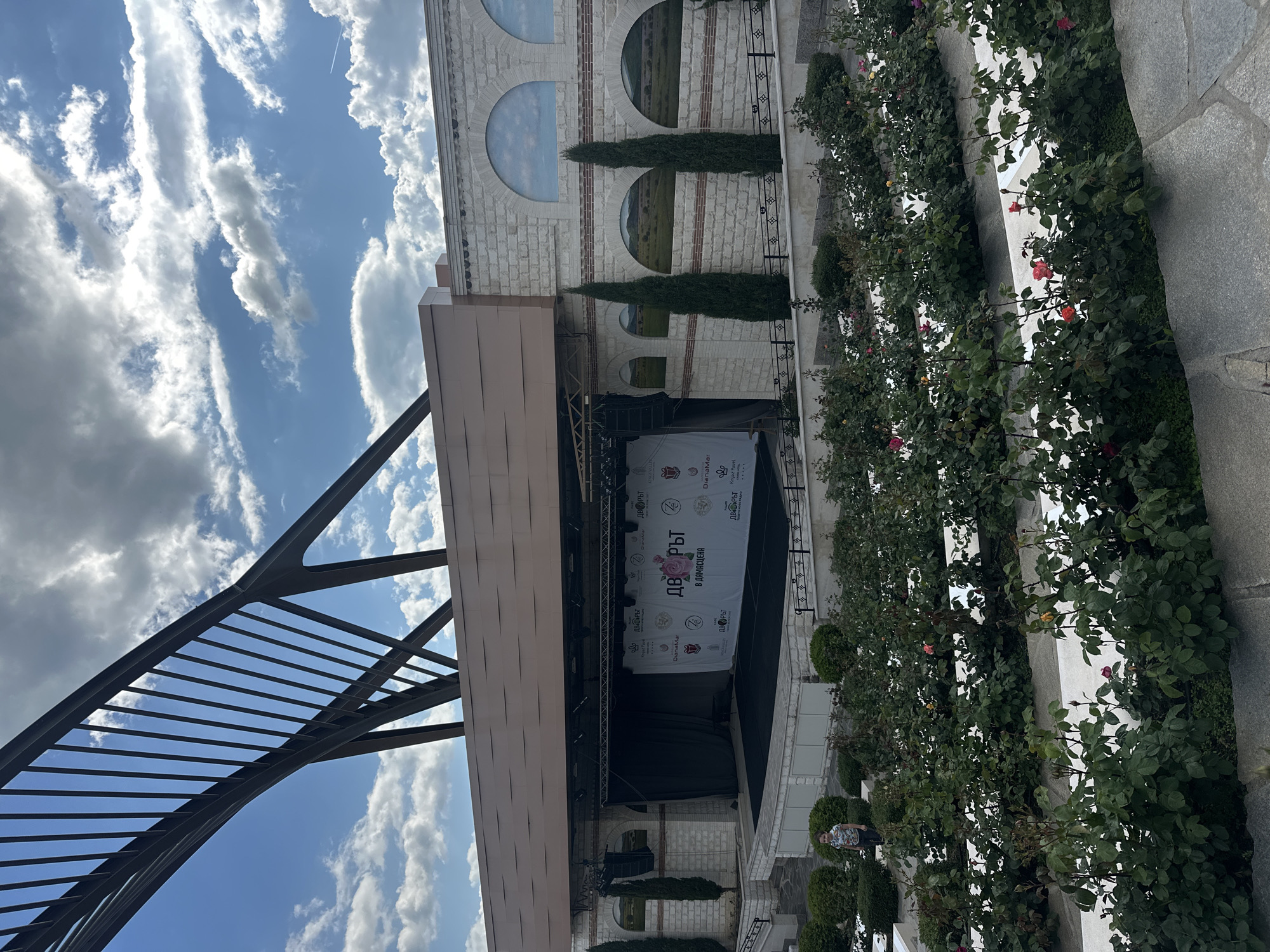 The image captures an outdoor event stage set against a stone building in what appears to be Skobelevo, Bulgaria. The stage features a banner with Cyrillic script, indicating local branding or sponsorship. Rose bushes line the area adjacent to the stage, adding a decorative element. An archway structure can be seen on the left side of the image. The architecture of the building includes arched windows, which are distinctive features. The sky is visible with light clouds. In the background, a landscape scene is visible through the arched windows. There are also trees planted near the building.