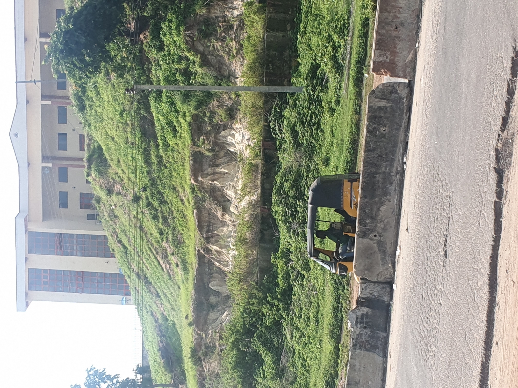 The image depicts a side view of a yellow and black auto-rickshaw, also known as a "keke," parked alongside a concrete barrier on a paved road. The auto-rickshaw has a clear windshield and a canopy, and a person can be seen inside, presumably the driver.
Behind the auto-rickshaw, a steep, rocky hillside is covered in lush green vegetation, with a prominent building visible on the left side of the frame. The building appears to be several stories high and has many windows with reddish-brown frames. The overall scene suggests an outdoor urban or semi-urban environment, possibly in a hilly area.
The lighting suggests it is daytime, with natural sunlight illuminating the scene. There are no strong indications of specific weather conditions, though the vibrant greenery implies it might be a time of year with sufficient rainfall. The image does not contain any visible text. The setting, with the auto-rickshaw and the building, aligns with the provided location context of Lokoja, Nigeria.