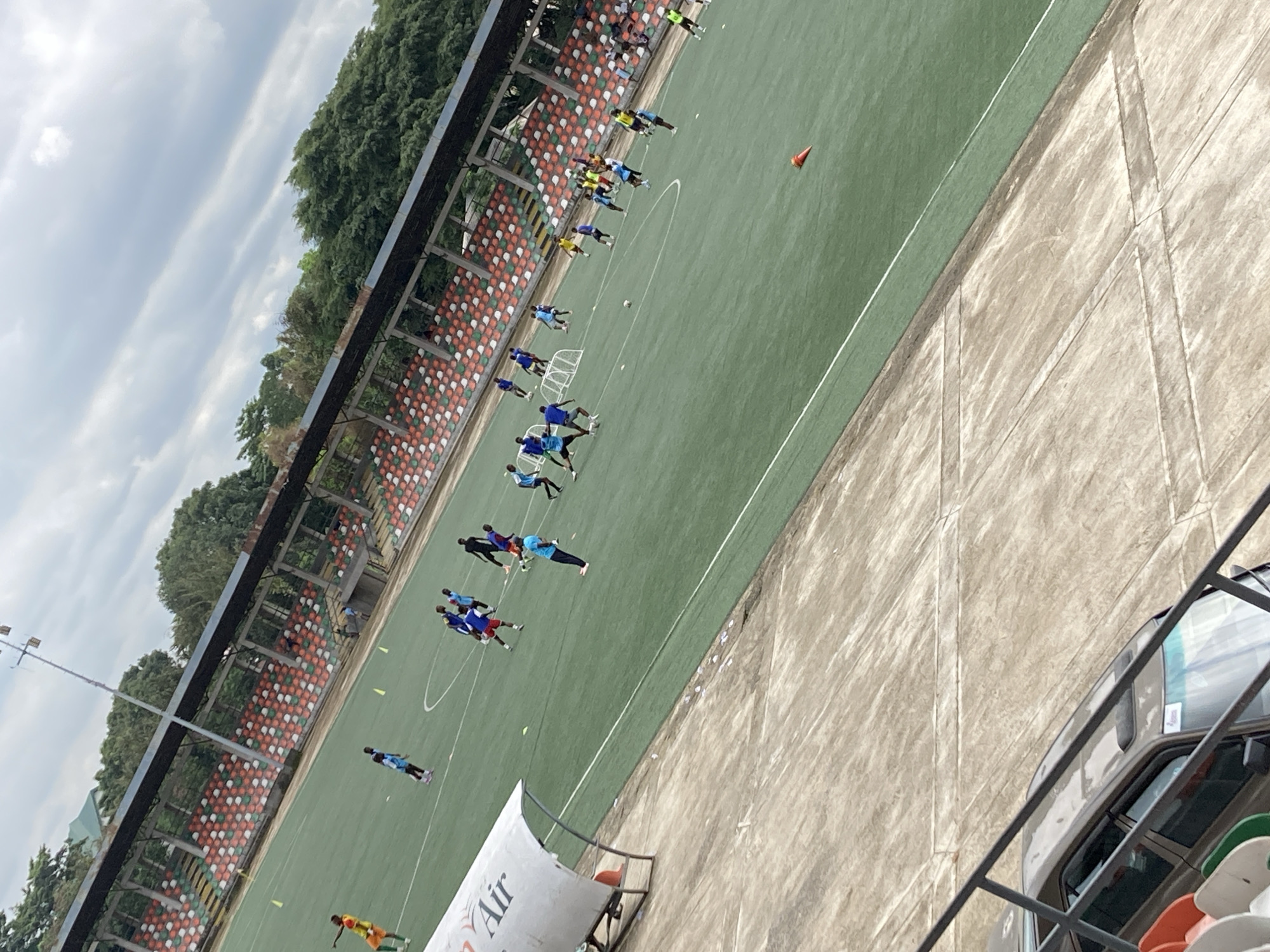 In Uyo, Nigeria, a football training session is underway at a stadium. The artificial turf field is occupied by numerous young players in various colored jerseys, engaged in drills and practice matches. A few observers are seated in the tiered stadium seating, which features a mix of orange, white, and green seats. A white canopy structure, possibly a dugout or shelter, is visible on the left side of the field. In the foreground, a concrete pathway slopes down towards a parked car. The sky above is overcast, with textured clouds.