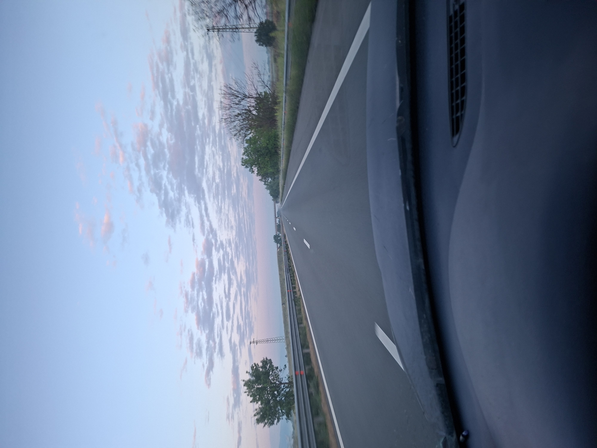 A view from inside a vehicle traveling on a multi-lane highway near Shishmantsi, Bulgaria. The highway has a white dividing line and guardrails on the left side. Trees and vegetation line the side of the highway. The sky is blue with clouds tinged with pink. The interior of the vehicle is visible on the right side of the image.