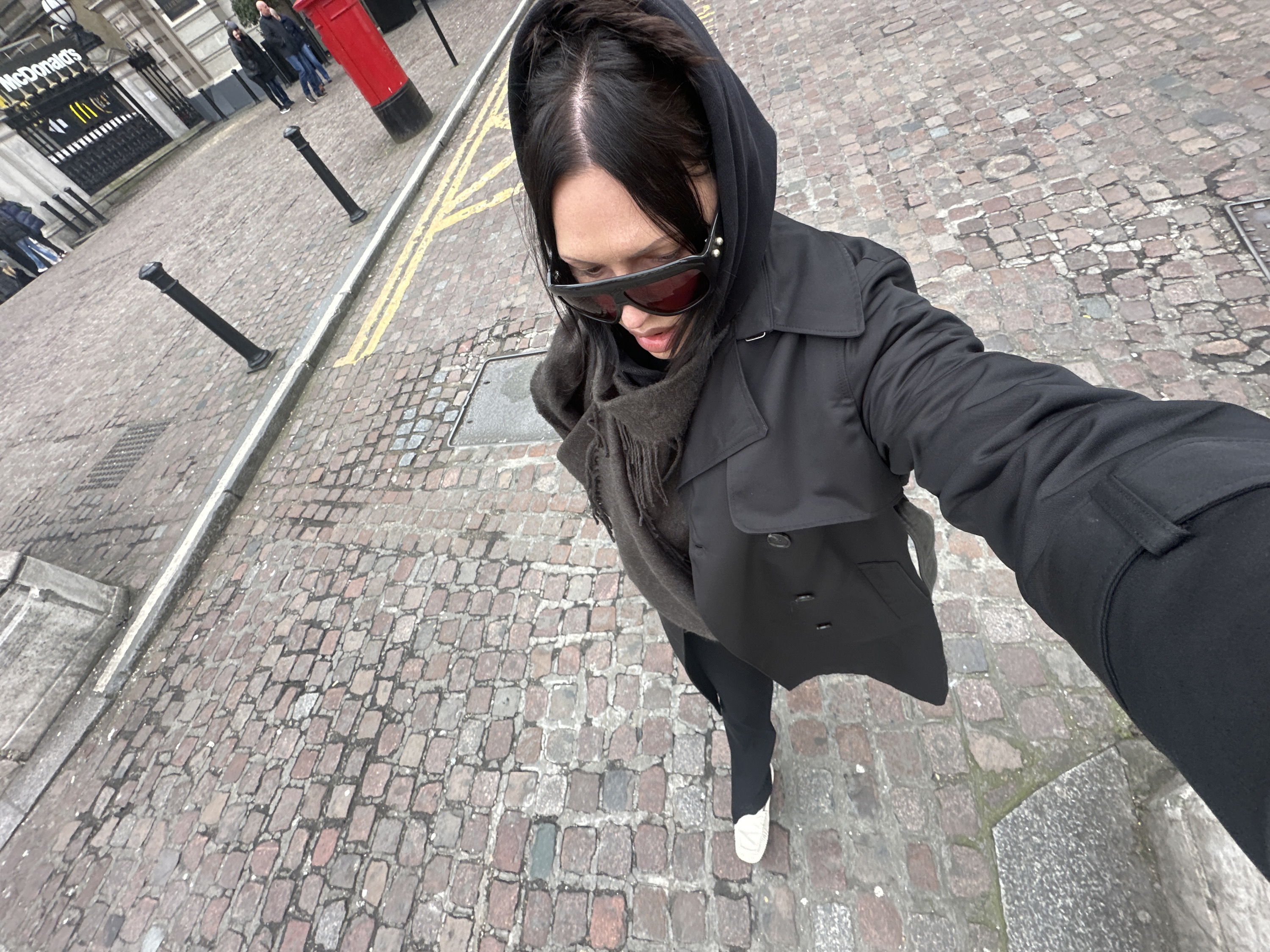 The image depicts an individual from an elevated, downward-angled perspective on a cobblestone street identified as Fleet Street, London, United Kingdom. The central subject, possessing dark hair, wears red-tinted sunglasses, a black hooded garment, a black trench coat, a dark grey scarf, black trousers, and white-soled footwear. Their right arm extends toward the viewer, clothed in black fabric. The street surface consists of irregular reddish-brown cobblestones, traversed by a grey kerb and double yellow lines. In the mid-ground right, a red cylindrical Royal Mail post box is present. Further background reveals a McDonald's establishment with its distinctive yellow 'M' logo and name, positioned behind black decorative metalwork gates on a stone building façade. Multiple black bollards are arranged along the street edge. Other pedestrians are visible in the distant background. The scene is an urban outdoor setting under ambient daylight.