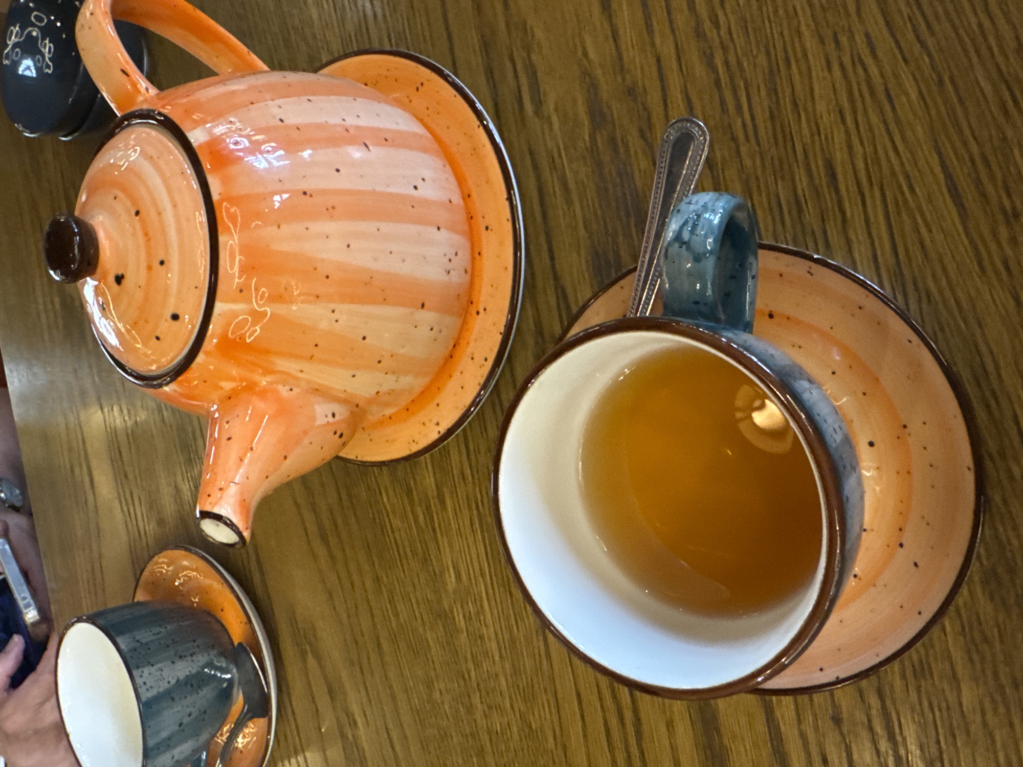 The image shows a close-up of a table with tea service. A vibrant orange and white striped teapot with black speckles sits on an orange saucer. Next to it is a dark blue cup and saucer, also speckled. The cup is filled with amber-colored tea, and a silver spoon rests on its rim. In the background, a person's hands are visible holding a smartphone, suggesting the scene is set in a cafe or restaurant. The table surface appears to be made of wood. The image does not contain any information about the location in the city of Veliky Novgorod, Russia.