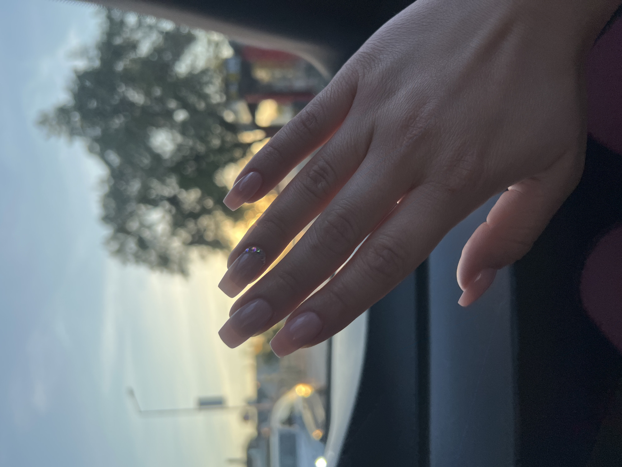 The image presents a close-up of a person's right hand, positioned with the palm slightly upward and fingers gently curled. The hand has light skin and features well-manicured nails, which are long, square-shaped, and painted in a translucent nude or light pink color. A small cluster of iridescent rhinestones adorns the ring finger near the cuticle.
The photograph is taken from inside a vehicle, looking out through a window. Dark, blurred elements of the car's interior are visible in the foreground and along the right side. The background, viewed through the window, is heavily out of focus, creating a soft bokeh effect. This blurred outdoor scene shows the silhouette of leafy trees against a sky that transitions from light blue at the top to a warm, golden glow lower down, characteristic of either sunrise or sunset. Distant lights, possibly from streetlights or other vehicles, appear as soft, circular blurs.
The overall setting suggests an urban or suburban environment, likely Plovdiv, Bulgaria, during the "golden hour" due to the warm, ambient lighting. The hand appears relaxed, and no specific activity other than being photographed is depicted. There is no visible text in the image.