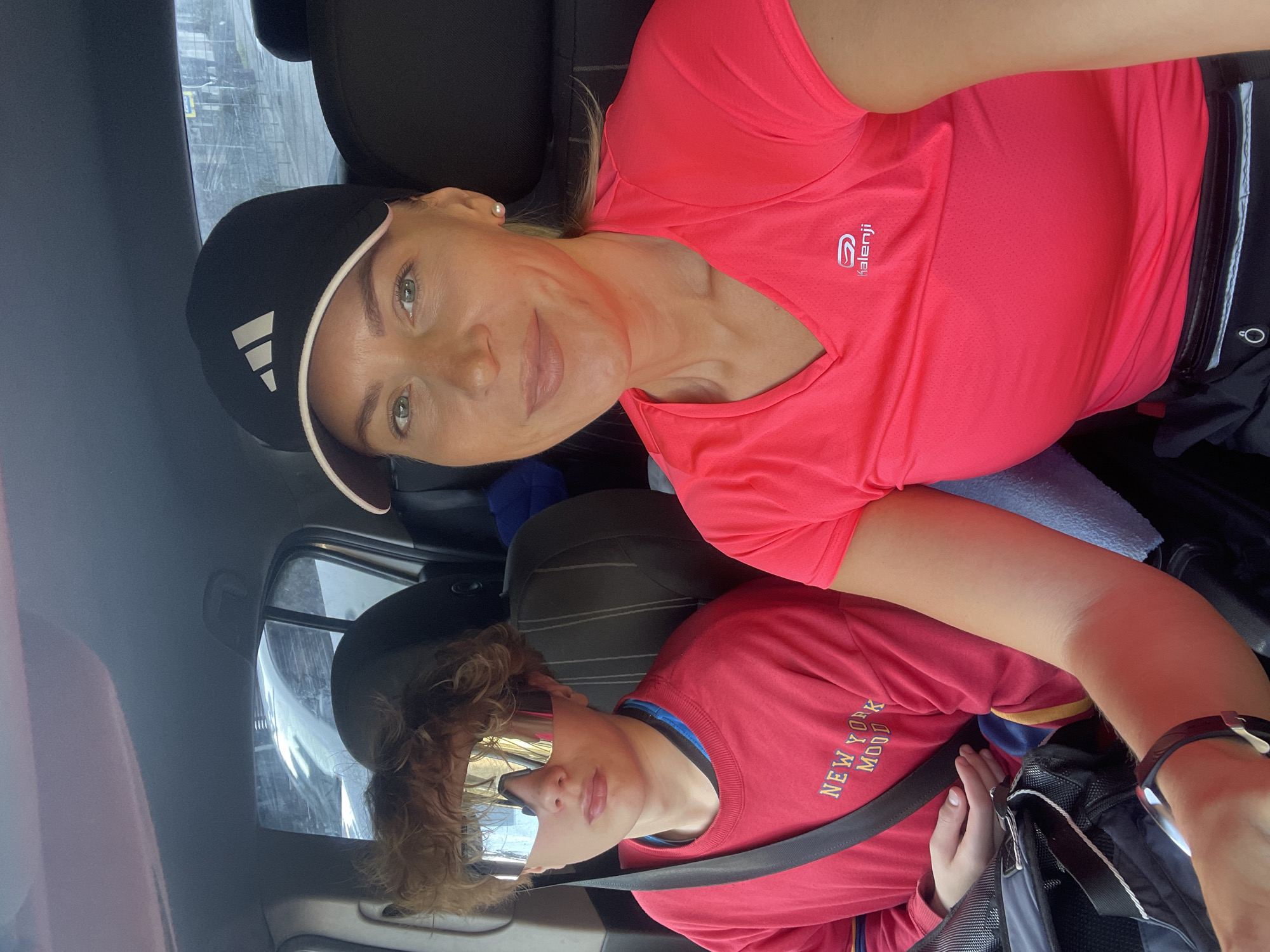 Two people are in a car. The woman in the passenger seat is wearing a pink Kalenji t-shirt and a black Adidas baseball cap with white stripes. She is smiling. The person in the back seat is wearing a red t-shirt with the words "NEW YORK MOOD" and reflective sunglasses. They are wearing a seatbelt. The photo was taken in Sochi, Russia, based on other available information.