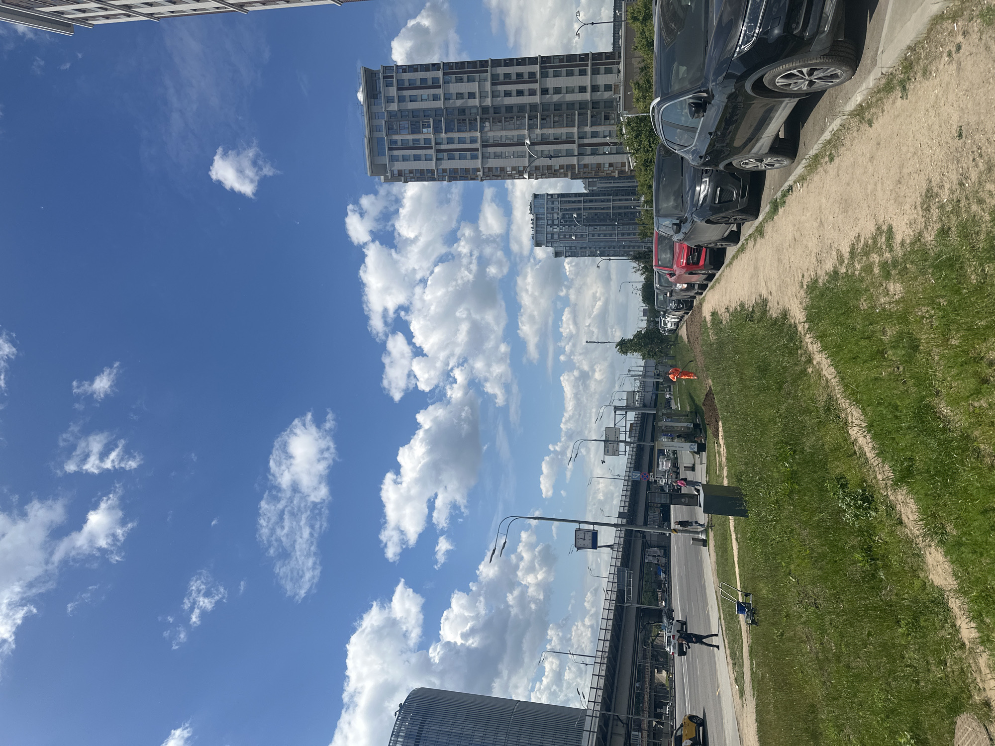 This media file captures an urban street scene in Moscow, Russia, under a clear, bright blue sky dotted with scattered white cumulus clouds. The image depicts a sunny daytime environment.
The scene features several modern, multi-story buildings, characteristic of a developed city. A prominent tall building with a repeating pattern of light and dark panels, punctuated by numerous windows, stands on the right side, extending upwards into the sky. Another similar building is visible further in the background. On the left side, a large cylindrical building with a ribbed exterior is partially visible.
A multi-lane road runs through the lower portion of the image, extending into the distance. Along the right side of the road, a line of cars is parked on a dirt shoulder. These include several dark-colored SUVs or crossovers, a red car, and a white car. Further down the road, a yellow car and other vehicles are visible.
Separating the road from a grassy area is a dirt path, with some worn tracks suggesting pedestrian or light vehicle traffic. The grassy area shows signs of green vegetation.
Several people are present in the scene. One individual in dark clothing appears to be walking on the left side of the road, pushing what looks like a shopping cart. In the distance, another person wearing an orange high-visibility vest is visible, possibly associated with construction or maintenance work. Overhead, an elevated structure, possibly a bridge or overpass, spans across the scene. Streetlights with square clock faces are mounted on poles along the road.
No legible text is visible on signs or buildings within the frame. The overall impression is one of a busy, modern urban landscape on a pleasant day.