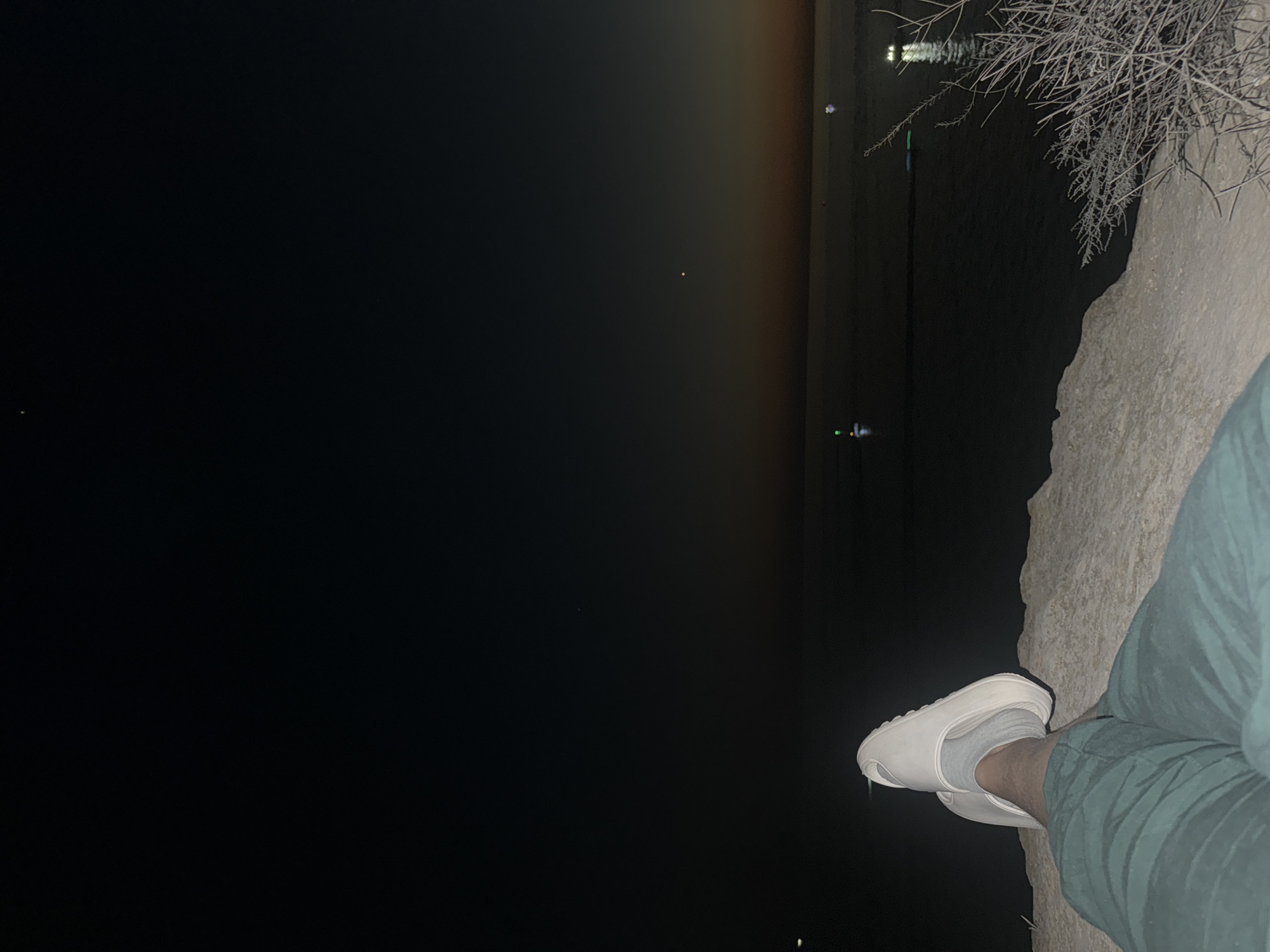 The image depicts a person's legs, clad in teal-colored pants and white slides with grey socks, resting on a rocky surface at night. The person is positioned in the foreground, facing away from the viewer and towards a body of water. The water reflects lights from distant objects, possibly boats or markers, creating a serene scene. The sky above is dark, transitioning to a faint gradient of orange and yellow near the horizon, suggesting either dusk or dawn. In the distance, the faint outline of what appears to be the city of Aktau, Kazakhstan, can be discerned along the shoreline. The overall atmosphere is calm and contemplative.
