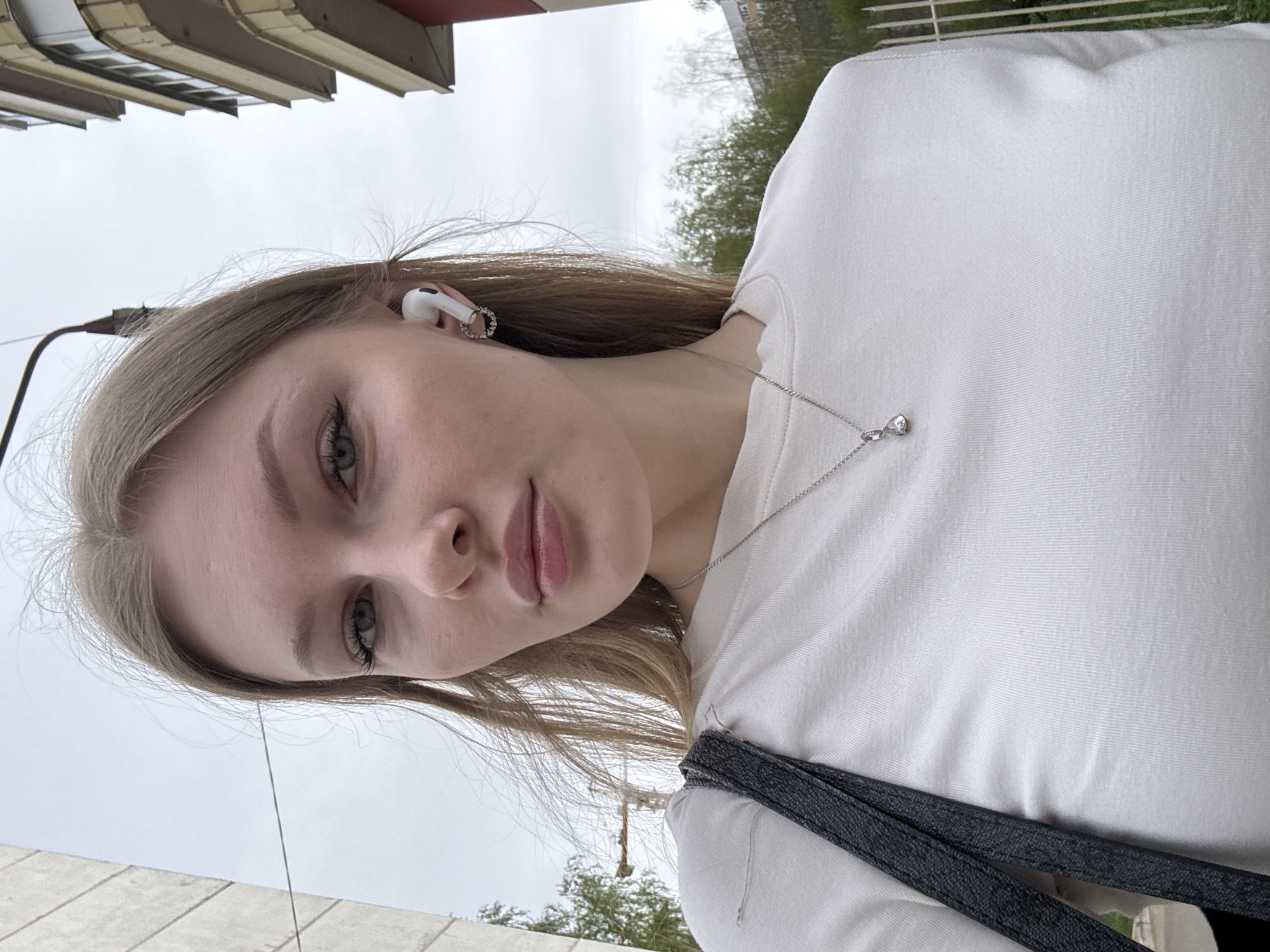 The image shows a selfie of a fair-skinned woman with light brown hair and green eyes. She is wearing a white long-sleeved shirt and a silver necklace with a small pendant. An earbud is visible in her left ear. She is outdoors, seemingly on a city street. Buildings and trees are visible in the background, with an overcast sky above. The perspective is angled upward. Based on the details provided and the visual characteristics, the location is specified as being in Perm, Russia.