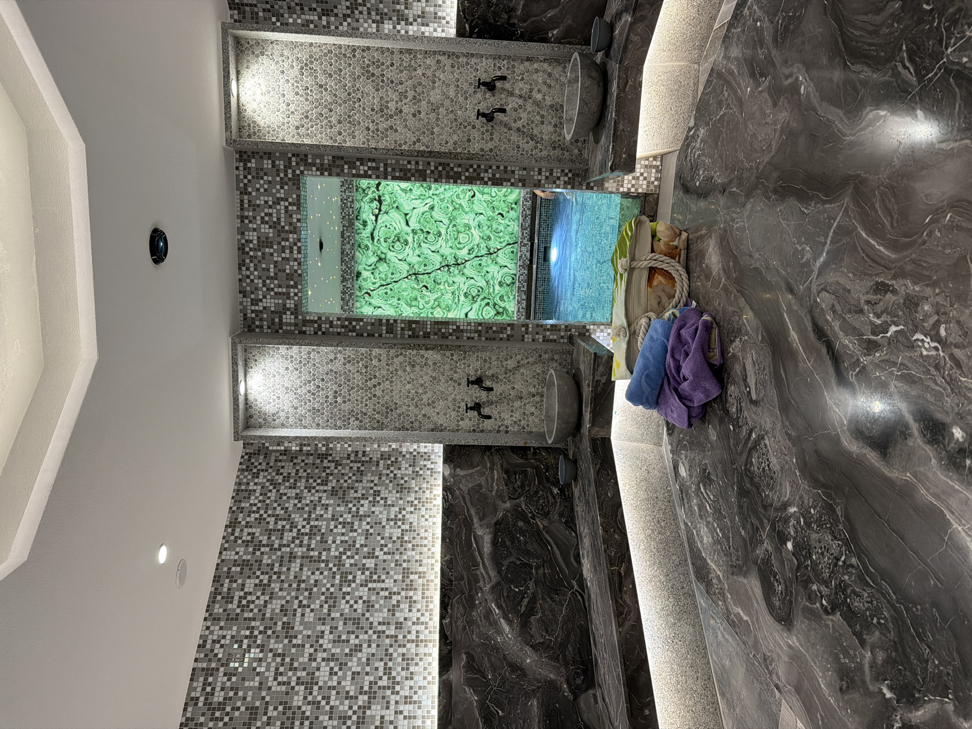 This media file depicts a luxurious indoor swimming pool area, possibly a spa or private residence, in Hisarya, Bulgaria. The scene features a small pool with clear blue water, illuminated from below, reflecting light onto the surrounding mosaic tile walls. One wall displays a large, vibrantly green malachite-patterned panel embedded with small lights, creating a starry effect.
Adjacent to the pool are two shower stalls, each with dark fixtures and stone basins. The area is finished with dark, veined marble countertops and walls, complemented by subtle, integrated lighting that casts a warm glow. On one of the marble surfaces, a straw tote bag rests, containing what appears to be a towel and possibly some personal items. Two folded towels, one blue and one purple, are placed atop the bag.
The overall ambiance is one of tranquility and opulence. The lighting suggests it is either evening or a dimly lit indoor space, enhancing the serene atmosphere. There are no people clearly visible, though the presence of towels and the inviting pool suggests recent or imminent use. The tiling patterns, varying from small mosaics to larger, polished marble, add texture and visual interest to the sophisticated design. The environment appears to be clean and well-maintained, exuding a sense of calm and relaxation.