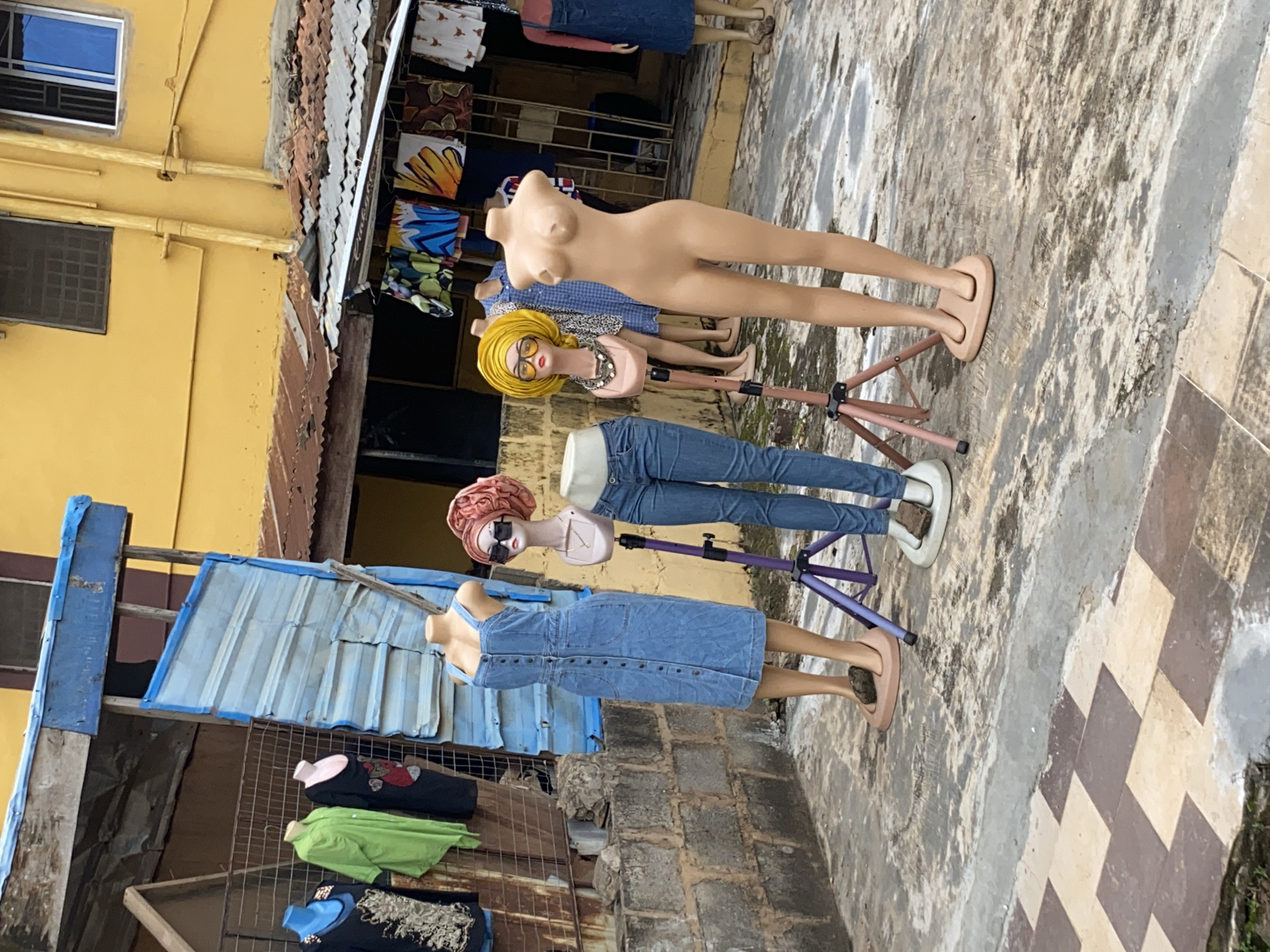 The image shows a row of mannequins on a street in Lagos, Nigeria. The mannequins are dressed in various items of clothing, including dresses, jeans, and tops. Some have wigs and accessories. They are standing on a paved area next to a yellow building and a corrugated metal structure. Hanging clothes are displayed in the background. The environment appears to be a marketplace or shopping area.