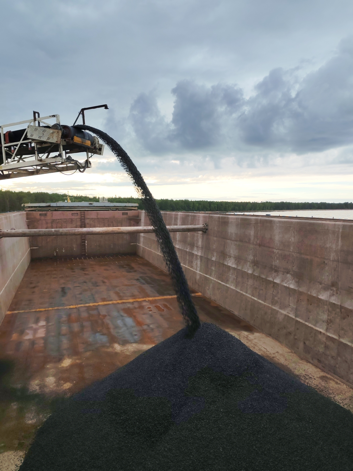 The image shows a barge being loaded with dark-colored gravel from a conveyor belt. The barge is a large, open vessel with weathered, reddish-brown metal walls. A significant pile of gravel has already accumulated in the foreground, with more material actively being discharged from the conveyor. The conveyor belt system is visible at the top left of the frame, extending over the barge. In the background, a body of water is visible, bordered by a dense line of trees under a cloudy, overcast sky. The scene appears to be an industrial loading operation, likely at a port or waterway near Shcheleiki, Russia. There are no people visible in the image.