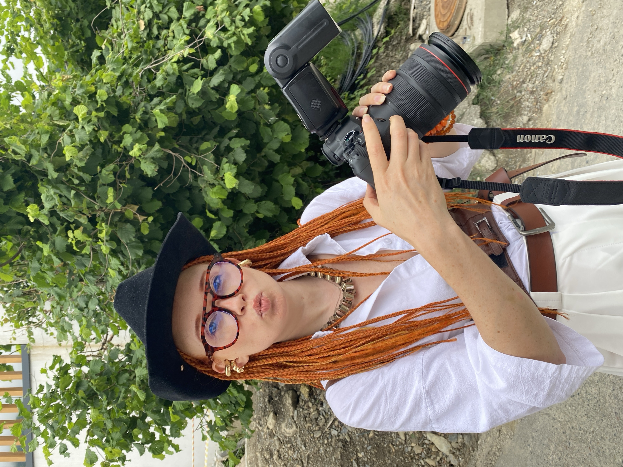 A woman with bright orange braided hair, wearing a black cowboy hat, large tortoiseshell glasses, a white button-up shirt, and white pants, poses for a photo outdoors. She is holding a professional Canon camera with a flash attachment, and has a brown leather belt and a distinctive chunky gold necklace. The background consists of lush green foliage and a gravelly area.
