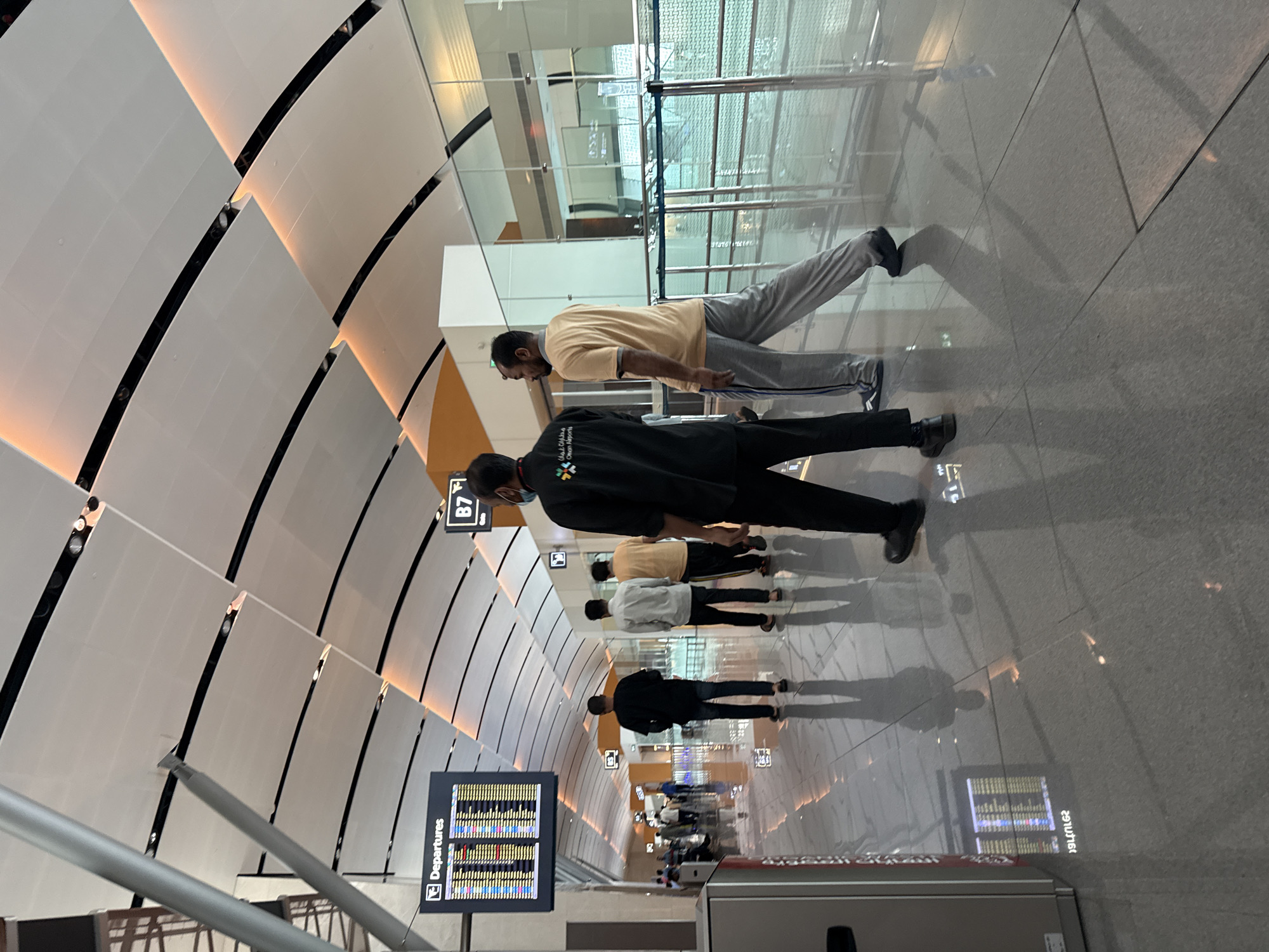 The image depicts an interior view of a modern airport terminal, likely in the city of Seeb Province, Oman, given the "Oman Airports" branding visible on the uniform of one of the individuals. The scene is characterized by a high, curved ceiling with integrated lighting, and polished, reflective floors that create distinct shadows of the people present.
In the foreground, two men are walking away from the camera. The man on the left wears a dark uniform with a colorful logo and text that appears to read "Oman Airports." He is facing slightly to the right. The man next to him wears a light yellow t-shirt and grey track pants with blue stripes, and is looking to his left.
Further into the terminal, several other individuals are visible, walking in various directions. To the left, a departures board displays flight information, with the word "Departures" clearly legible at the top. Another sign, labeled "B7," is visible further down the concourse, presumably indicating a gate number. The terminal appears to be a well-lit and spacious area, with large glass panels and barriers creating walkways. The overall impression is one of a busy but orderly airport environment.
