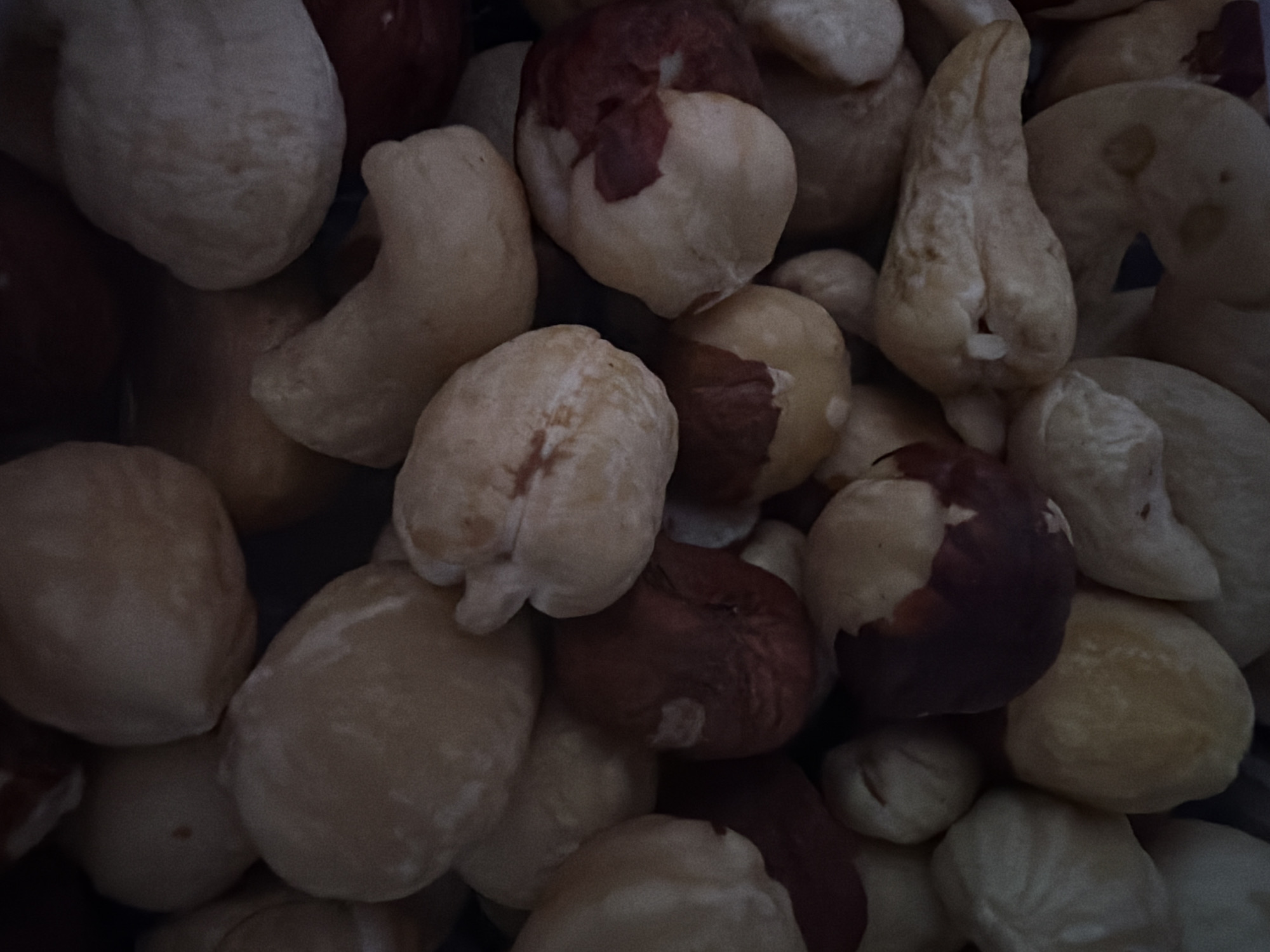 This media file is a close-up, overhead shot of a pile of hazelnuts. The nuts are mostly shelled, with some retaining a reddish-brown skin. The lighting is dim, casting shadows and highlighting the textures of the nuts. The background is dark, making the nuts the sole focus. There is no discernible text, people, or specific setting information. The activity depicted is simply a collection of nuts, suggesting a potential ingredient or food item. The image's overall tone is muted due to the low light conditions.