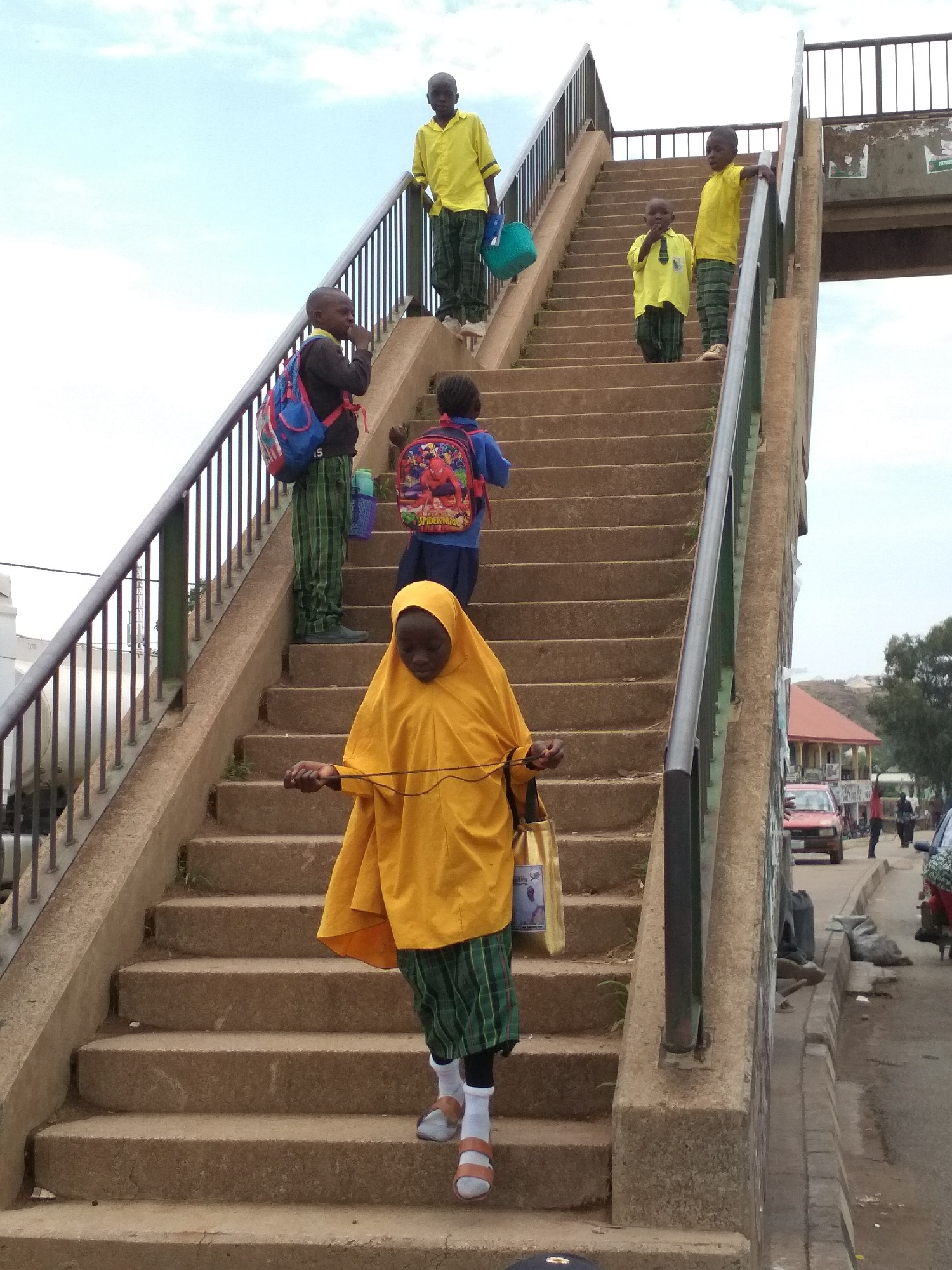 In Gura Top, Nigeria, a group of schoolchildren in yellow shirts and plaid pants ascend and descend a concrete overpass. One girl, wearing a yellow hijab, walks down the stairs with a bag and a jump rope. Two boys stand at the top of the stairs in yellow shirts and plaid pants. One boy has a blue bucket. Two other boys with backpacks are also on the stairs. The overpass has metal railings, and cars and pedestrians are visible on the street below.