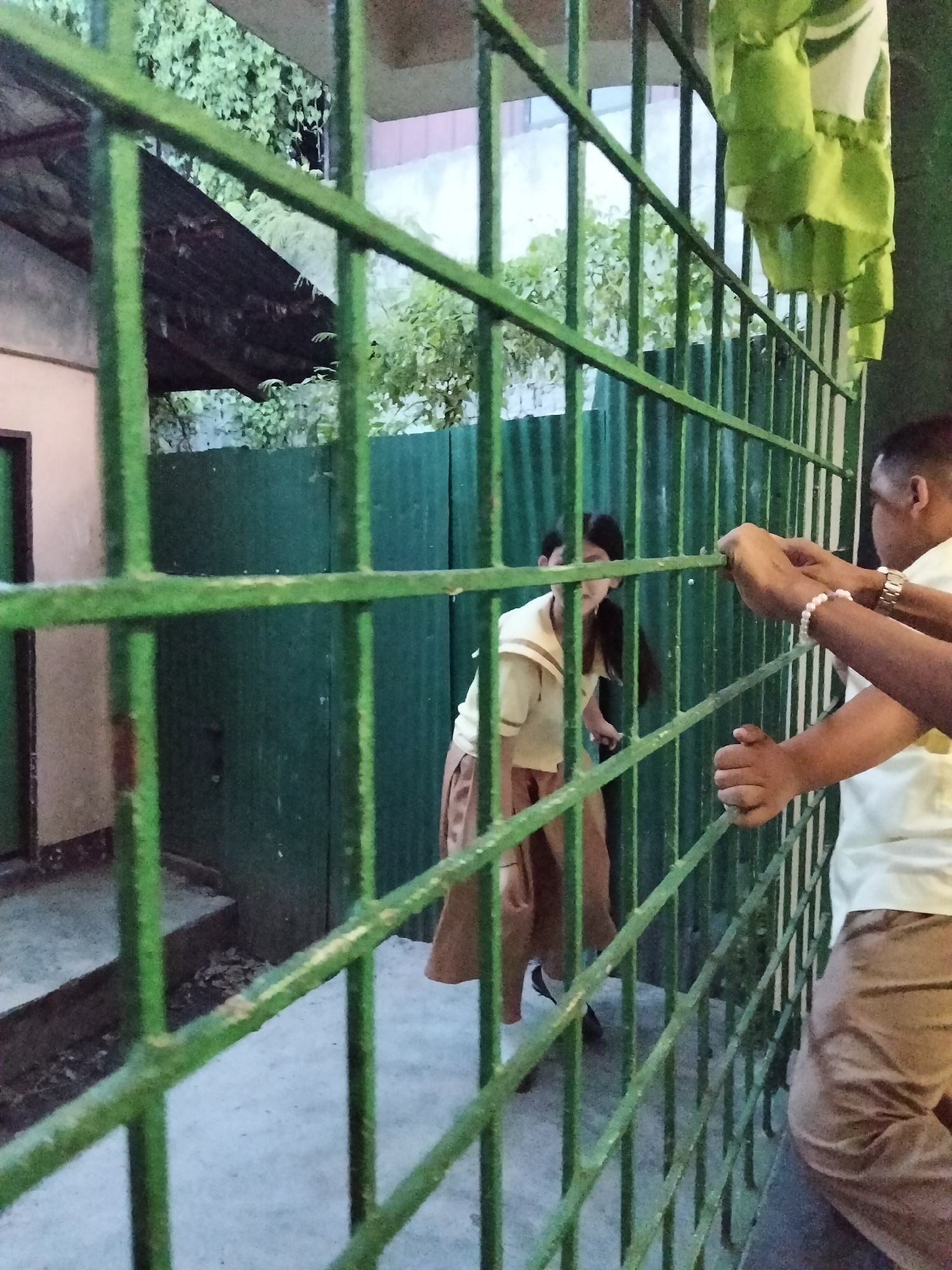 Two people are visible through a green metal gate. The person in the foreground, on the right, appears to be an adult male wearing a light-colored shirt and brown pants. He is holding onto the gate with both hands. His left wrist has a white beaded bracelet and a watch. The second person, in the background and to the left, is a younger individual, possibly a student, wearing a light-colored school uniform with a pleated skirt. This person is looking towards the gate, their expression somewhat obscured.
The setting appears to be an outdoor area with a concrete floor. Behind the gate, there is a green corrugated metal fence. To the left, a portion of a building with a brown roof and a door with a dark frame is visible. Overgrown greenery and vines can be seen on top of the roof. In the background, there is a light-colored wall and more foliage. The overall lighting suggests it is either early morning or late afternoon, with a diffused light. The location context of Davao City, Philippines, might imply a warm climate. The scene gives a sense of interaction or observation through the barrier. No text is visible in the image.