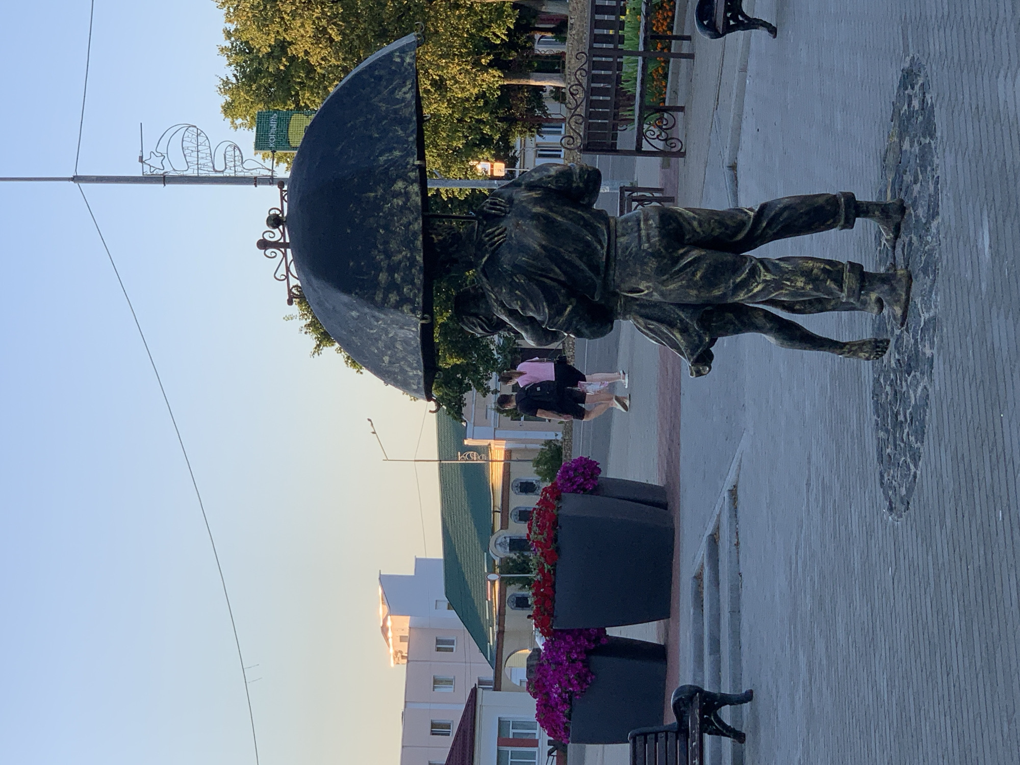 The image depicts a street scene in the city of Kapyĺ, Belarus, featuring a bronze sculpture of a couple embracing under an umbrella. The sculpture is positioned in the foreground, with the man holding the woman in a dance-like pose. Behind the sculpture, large planters filled with vibrant purple and red flowers are visible, adding a splash of color to the paved walkway.
In the middle ground, two individuals, a man and a woman, are walking away from the camera. The woman wears a pink top and dark shorts, while the man has a black t-shirt and shorts. Further in the background, several buildings are present, including one with a green roof and another with a light-colored facade. The sky above is clear and blue, suggesting it is daytime. Benches are strategically placed along the walkway, offering seating for visitors. The overall impression is a well-maintained public space, likely a park or town square, designed for relaxation and enjoyment. The lighting suggests it is either early morning or late afternoon.