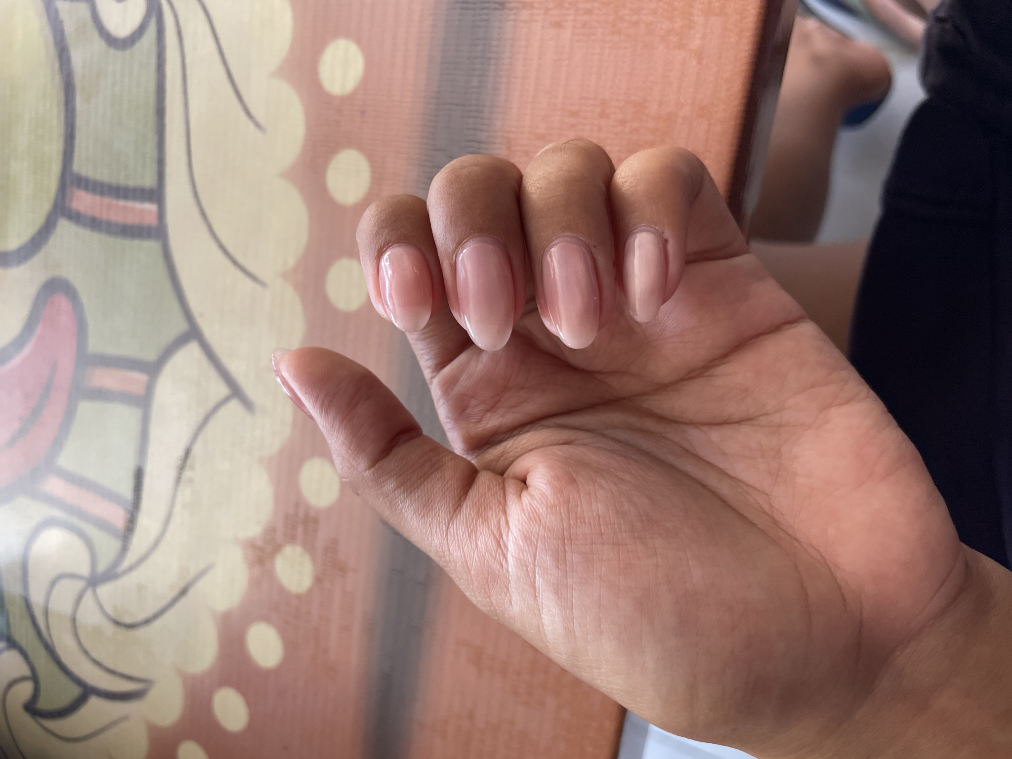 A close-up, overhead shot shows a person's left hand, with their fingers gently curled. The nails are short and oval-shaped, painted with a sheer, pale pink polish. The skin on the hand is smooth and a medium brown tone, with visible lines on the palm and fingers. The hand rests on a surface that appears to be a wooden table with a patterned overlay. The overlay has a cream-colored background with scattered yellow dots and a decorative border featuring abstract, swirling lines in green, black, and pink. The background is out of focus, but suggests an indoor setting. The image does not provide any information to determine the city of Denpasar, Indonesia.